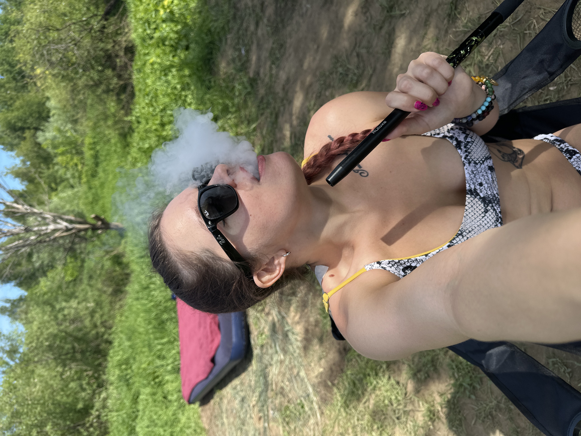 This image captures a female subject outdoors in Ladoga, Russia, on a bright, sunny day. The subject, positioned centrally, is taking a selfie, holding a black cylindrical hookah hose labeled "RULUXE" in her left hand (viewer's right) and exhaling a significant plume of white vapor. She wears black Ray-Ban sunglasses and a two-piece bikini with a black and white snakeskin pattern and yellow trim. Her dark hair is pulled back, with a braided section visible over her right shoulder (viewer's left) interwoven with a yellow element.
Her visible accessories include small silver hoop earrings and multiple beaded bracelets on her left wrist, featuring colors such as turquoise, black, yellow, and green. Her fingernails are painted bright pink. A mole is present on her right shoulder (viewer's left), and faint tattoos are visible on her upper left chest/shoulder area and lower abdomen. She is seated in a black folding chair, parts of which are visible in the foreground.
The background consists of a natural outdoor setting with vibrant green grass and dense foliage from trees and bushes under a clear blue sky. To the far left, a dark blue inflatable mattress covered with a red blanket or towel rests on the ground. The ground beneath the subject is a mix of dry grass and patches of exposed soil. The lighting indicates direct sunlight.
