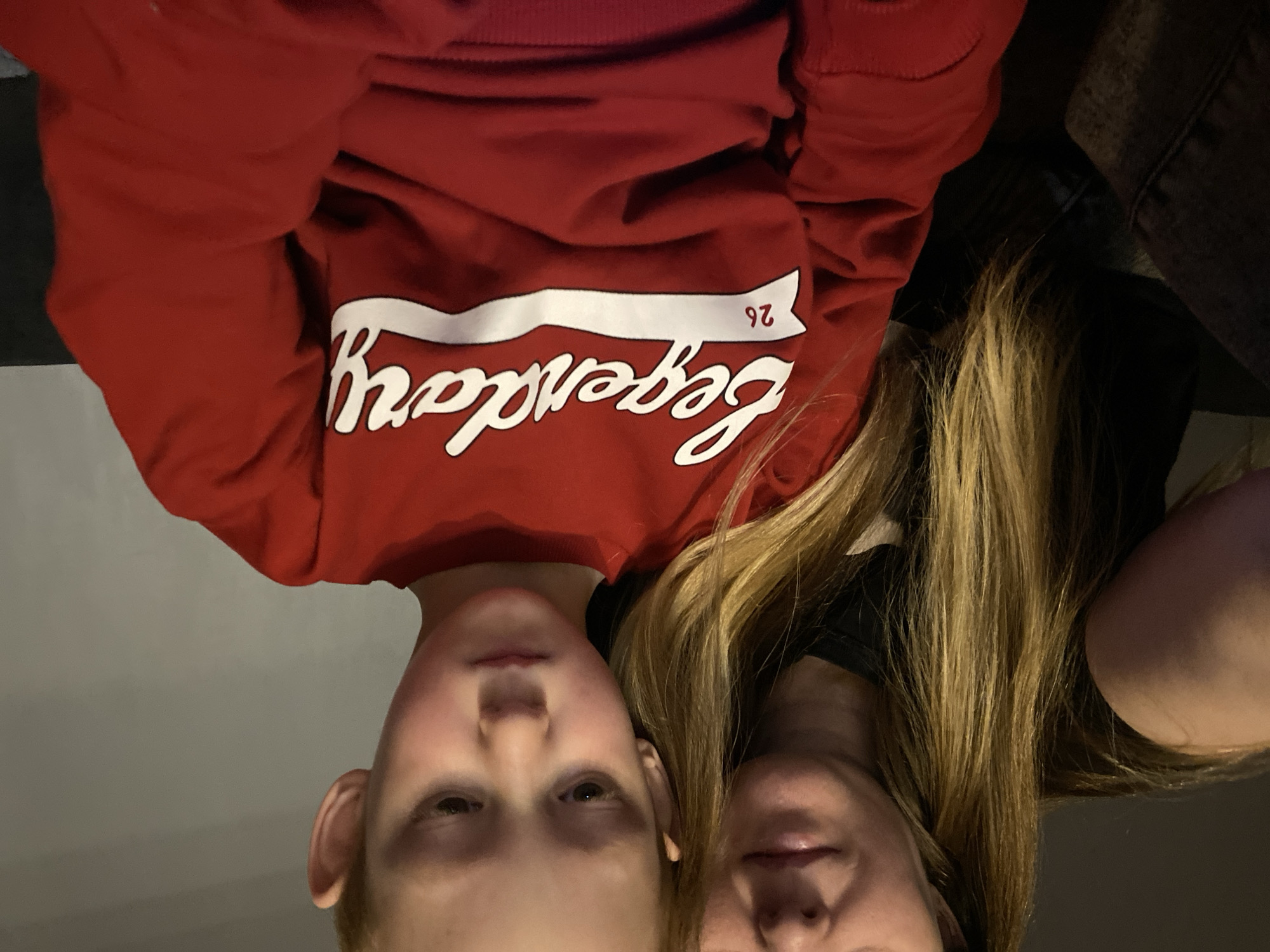 Two individuals, a child wearing a red sweatshirt with the word "legendary" printed on it, and an adult with long blonde hair, are positioned closely together, facing the camera. The image is rotated 180 degrees. The background is indistinct. No location in the city of Noviy, Russia, is apparent.