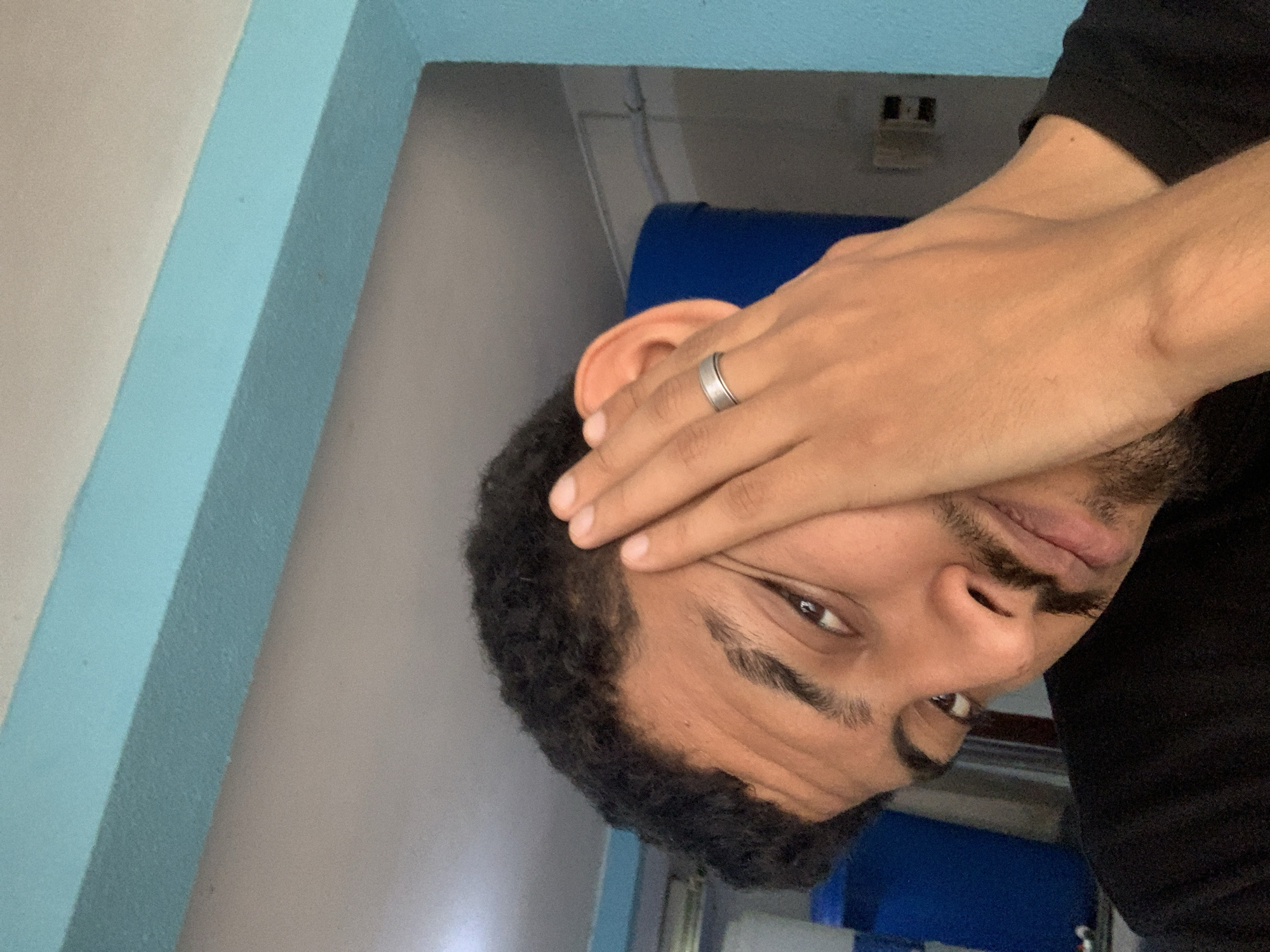 An eye-level shot captures a person, likely male, with medium-dark skin, dark curly hair, and a beard or stubble, in an indoor setting in Tacarigua, Venezuela. The person's left hand is partially covering their face, with fingers resting on their forehead and upper cheek, obscuring part of their right eye. A silver-colored ring is visible on their left ring finger. Their visible eye, which is dark brown, gazes directly towards the viewer. The person is wearing a black long-sleeved garment, possibly a shirt or sweater.
The background features distinct architectural elements. A prominent light blue beam or frame runs diagonally through the composition, contrasting with walls that appear to be light gray or beige. A darker blue object, possibly furniture or another structural detail, is also visible in the background, along with faint hints of what might be a light fixture or vent in the upper area. The image is significantly tilted, giving the impression that the subject and background are rotated. No text is visible in the image.