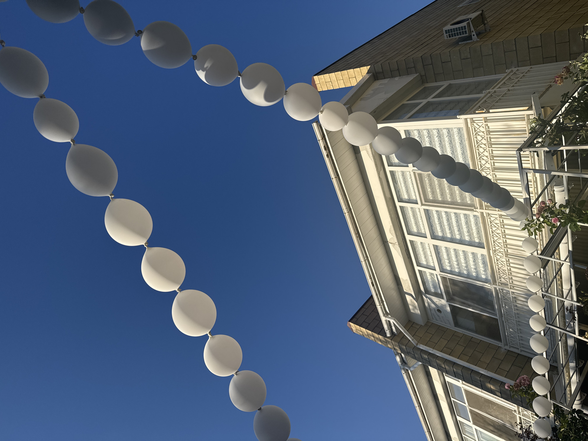 A string of white and gray balloons hangs in a clear blue sky above a multi-story building in Sary-Oy, Kyrgyzstan. The building, constructed with light-colored brick and white trim, features multiple windows, some with patterned blinds. A balcony with a decorative white railing is visible on the second floor. An outdoor air conditioning unit is mounted on the exterior wall. Greenery, including rose bushes, can be seen at the base of the building. The balloons are arranged in two separate strings, forming arcs against the sky.