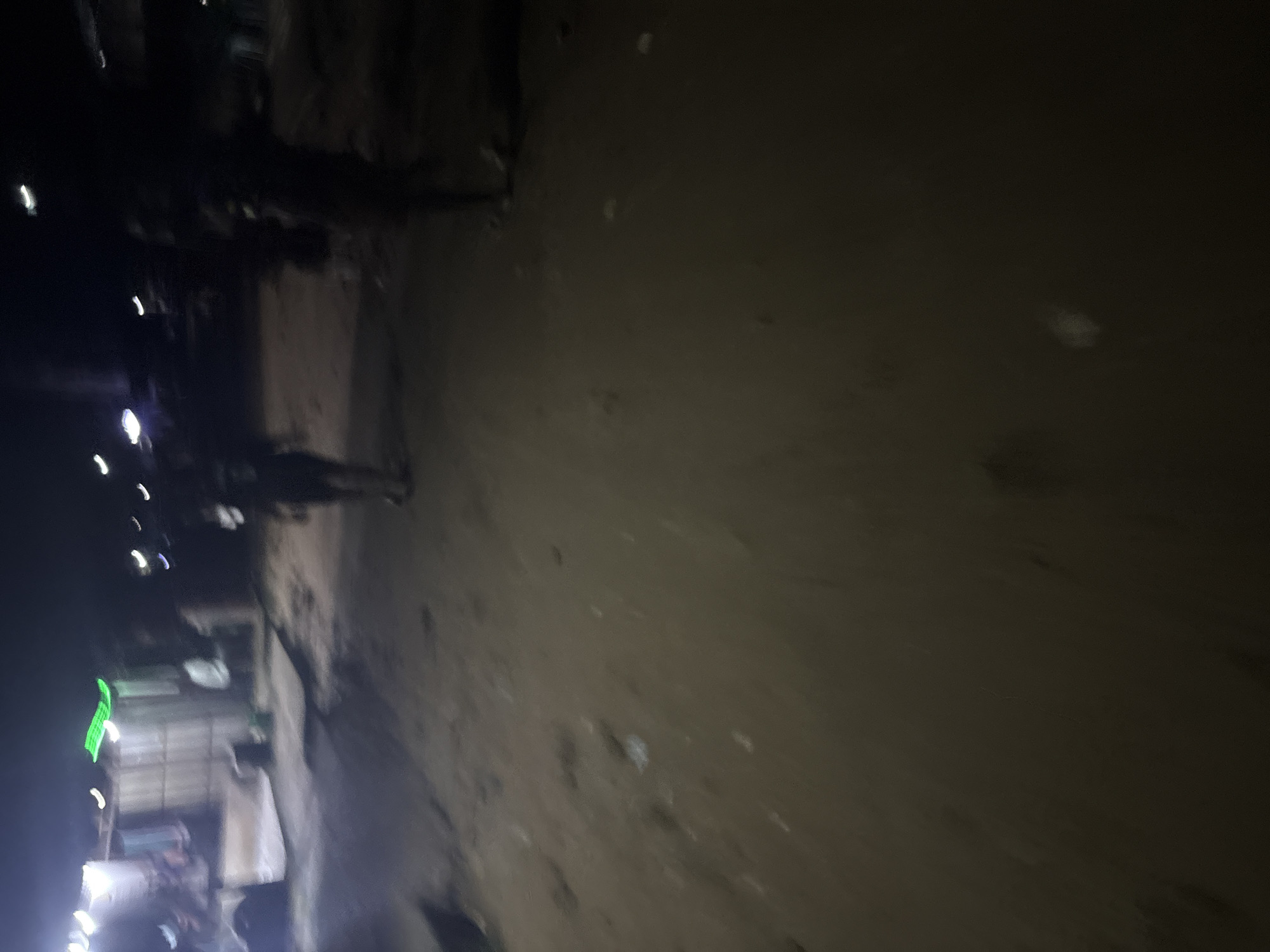 The image depicts a dimly lit outdoor scene, likely at night, on a sandy or dirt ground. In the background, a structure resembling a stall or shop with corrugated metal walls and a roof is visible, illuminated by artificial lights. There is a bright green illuminated sign on the roof of this structure. Several individuals are present. One person, silhouetted and appearing to stand, is in the mid-ground, facing away from the camera. To the left of this person, another figure is partially visible, also standing. Further left, near the structure, a person in a light-colored shirt is standing. In the far background, more indistinct figures and structures are visible, suggesting an area with some activity. The ground is uneven and littered with small debris. The overall impression is of a casual outdoor gathering or market area at night, possibly in the city of Onitsha, Nigeria, given the context.