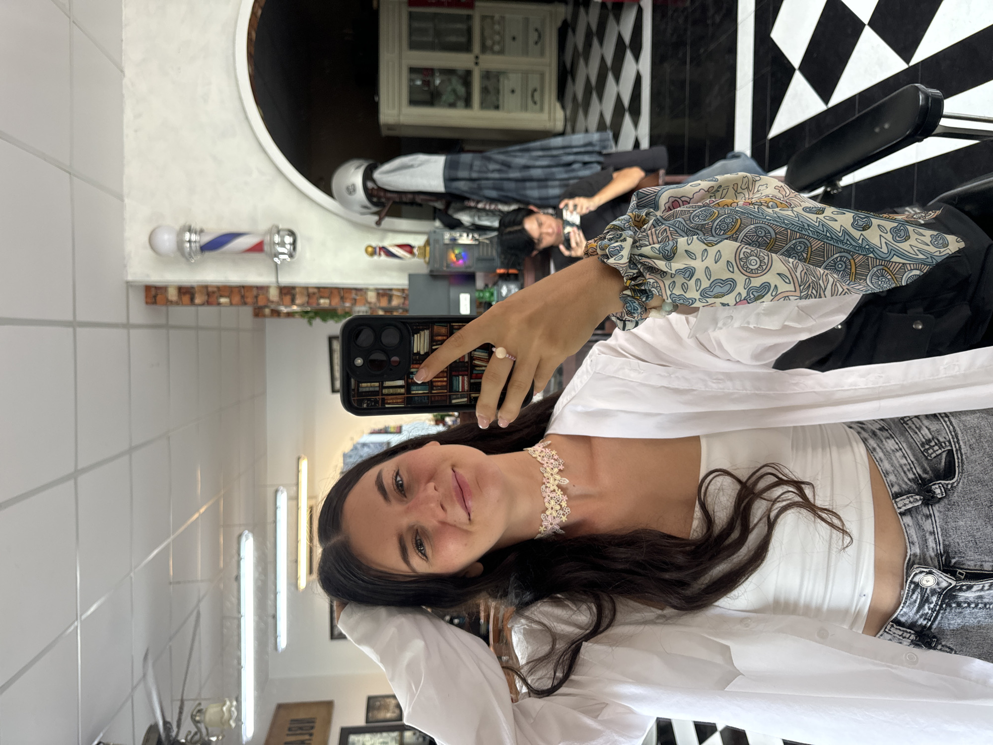 A young woman with long dark hair is taking a selfie in a barber shop. She is wearing a white tube top, a white open button-up shirt, and distressed denim jeans. She has a light pink and yellow floral choker necklace and a ring on her right index finger. Her left hand is touching her hair, and her right hand is holding a phone with a book-patterned case. A colorful patterned scrunchie is on her right wrist. In the background, a barber pole with red, white, and blue stripes is visible. A man with dark hair is sitting in a barber chair in the background, looking at his phone. The floor has a black and white checkered pattern. The barber shop appears to be located in the city of Phan Thiết, Vietnam, as indicated by the Vietnamese writing on a sign in the background.