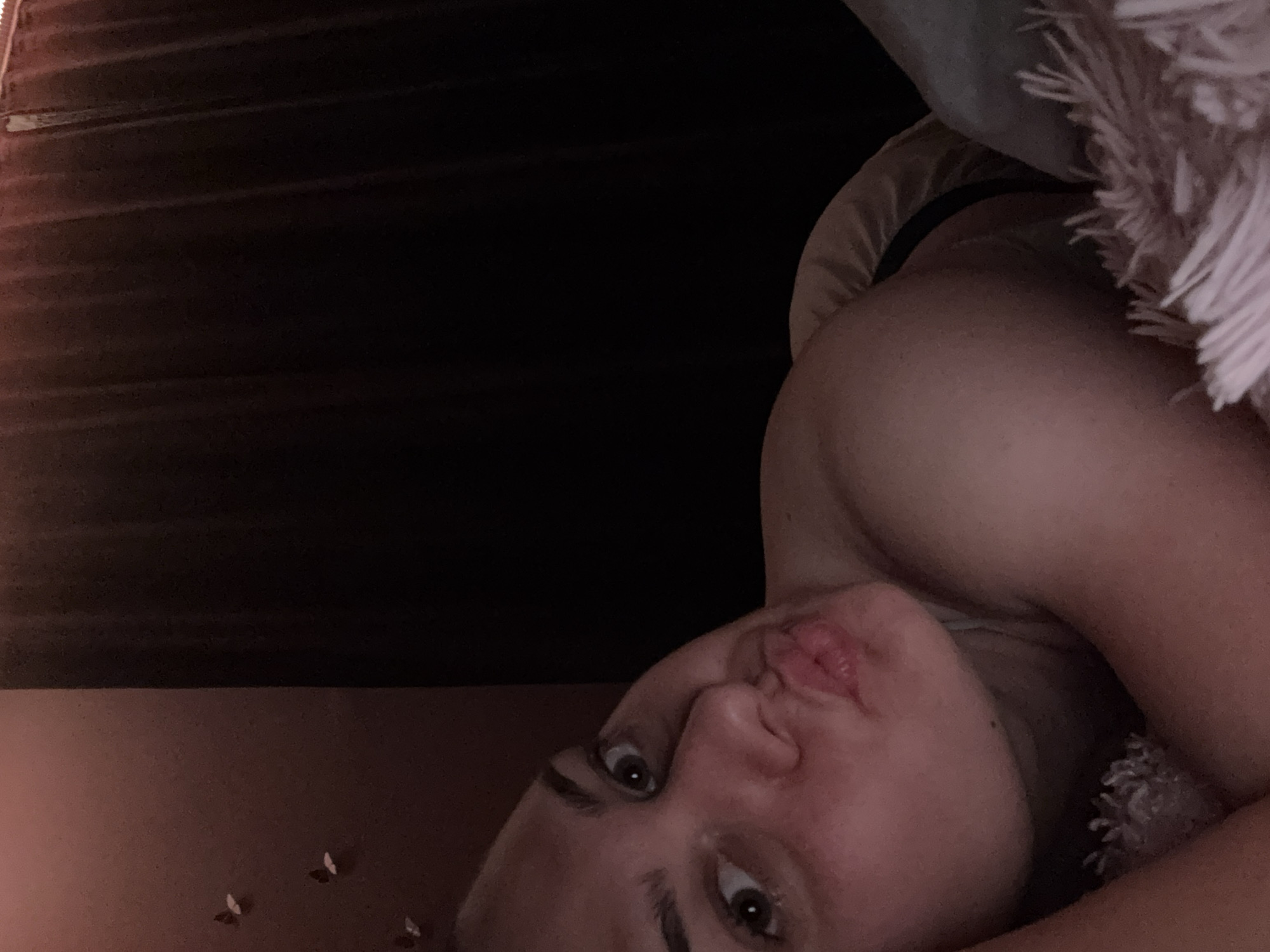 The image shows a close-up, waist-up view of a young woman lying down, facing the camera and puckering her lips. She has light skin and brown eyes, with her hair pulled back. She is wearing a dark-colored garment that is partially visible. The background is dark and out of focus, with what appears to be dark curtains or drapes. To the left of the woman, on a reddish-brown wall, there are several small, decorative butterfly cutouts. The woman is positioned on what looks like a bed with light-colored bedding and a pink, fluffy throw blanket in the foreground. The lighting is dim and warm, creating a soft glow on the woman's face and upper body. There is no indication of the specific location being in the city of Rogosh, Bulgaria, or any other city.
