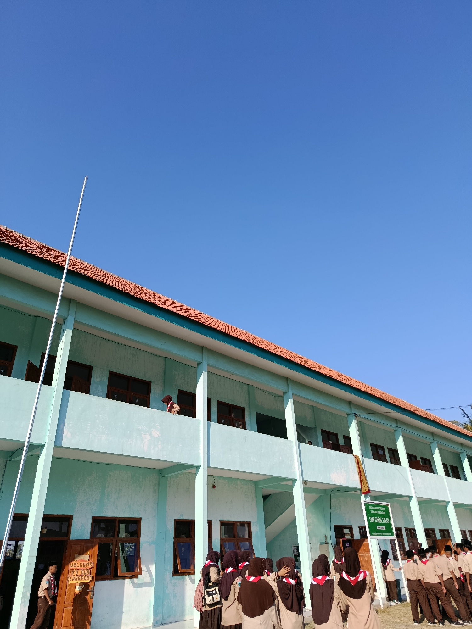The image depicts a school building with a pale blue facade and a terracotta-tiled roof. The building has multiple floors with balconies and dark-framed windows. A flagpole stands tall to the left of the building, extending into a clear blue sky. A group of students, dressed in uniforms that include brown shirts, dark pants or skirts, and red and white neckerchiefs, are gathered in front of the building and on the upper floor balconies. Some students appear to be standing in formation, while others are interacting. A wooden door with signage reading "WELCOME TO OUR CLASSROOM" is visible on the ground floor. A green sign with text in Indonesian, "SMP DARUL FALAH," is placed near the entrance. The setting appears to be a sunny day in Ramban Wetan, Indonesia, as indicated by the architectural style, the students' attire, and the signage.