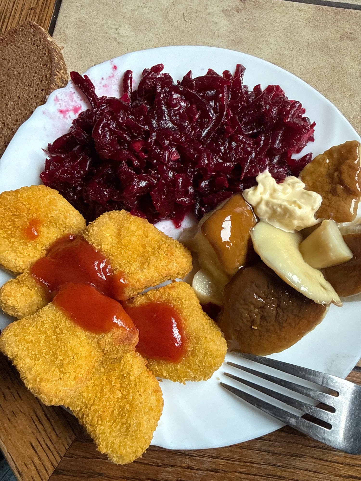 A white plate with food rests on a wooden surface. The plate contains shredded beets, breaded chicken nuggets drizzled with ketchup, and sliced mushrooms with a dollop of mayonnaise. A fork is positioned on the lower right side of the plate. The wooden surface appears to be a table. The image is cropped from a vertical perspective. There are no people present in the image. The food is presented in a home-style setting and does not provide any specific indications of its location within the city of Galinė, Lithuania.