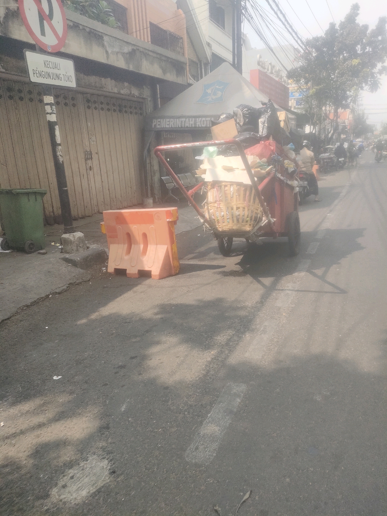 The image depicts a street scene in Surabaya, Indonesia. In the foreground, a garbage cart laden with trash is positioned on the right side of the road. The cart appears to be made of red metal with a woven basket attached. Behind the cart, a person is seated on a motorcycle. To the left of the garbage cart, there's an orange traffic barrier. Further left, a green trash bin with wheels is visible. A street sign indicating "KECUALI PENGUNJUNG TOKO" (Except Shop Visitors) and a no-parking symbol are mounted on a pole. In the background, buildings line the street, and various vehicles, including motorcycles, are present, suggesting traffic activity. The overall atmosphere appears hazy, possibly due to pollution or weather conditions. The area seems to be a commercial or mixed-use zone within the city.