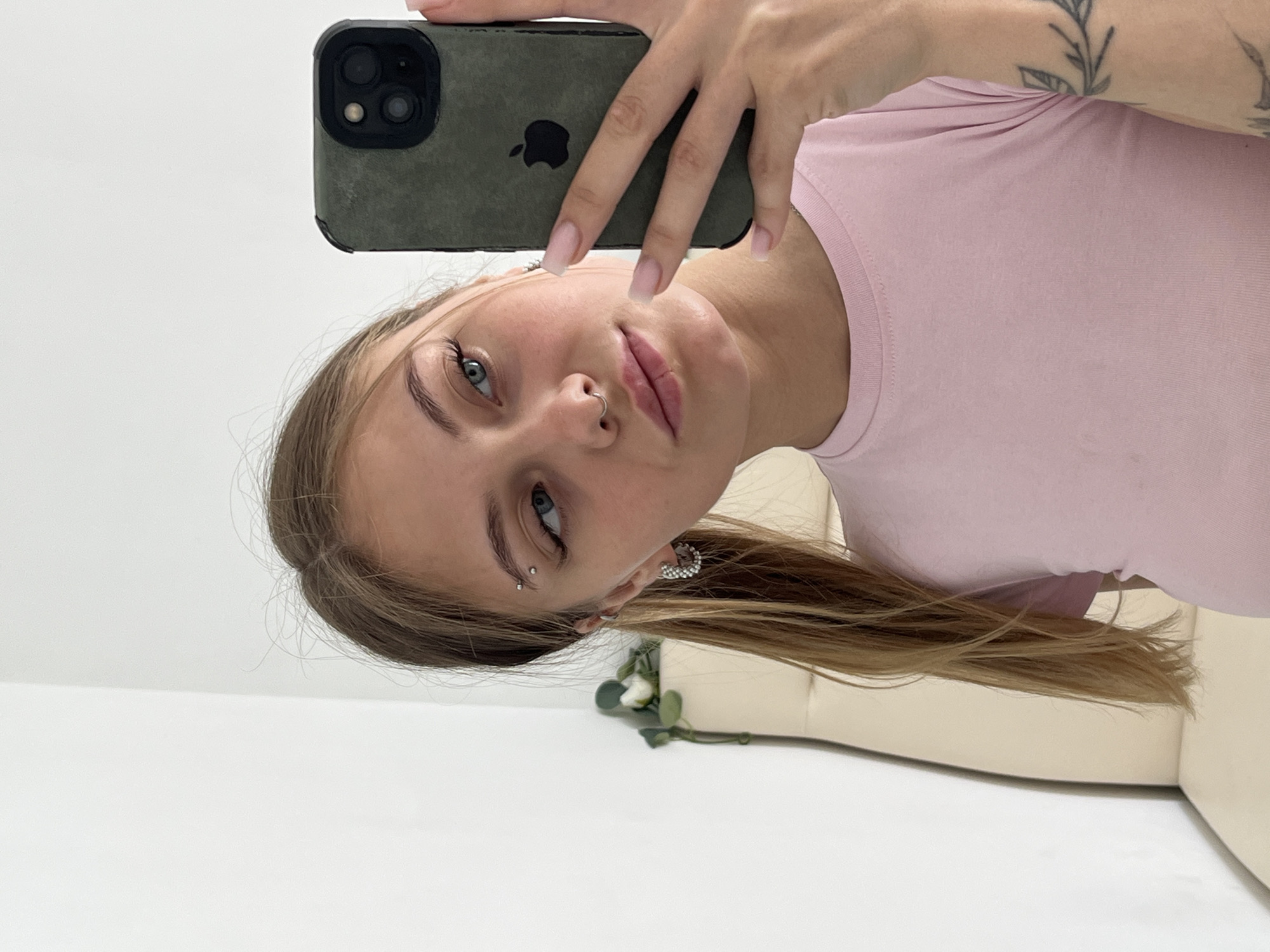 A young woman is taking a selfie with a smartphone. She has fair skin, light brown hair, and blue eyes. She is wearing a pink shirt and has a nose ring and an eyebrow piercing. A tattoo is visible on her upper arm. The background appears to be a room with white walls. The specific location is not discernible from the image.