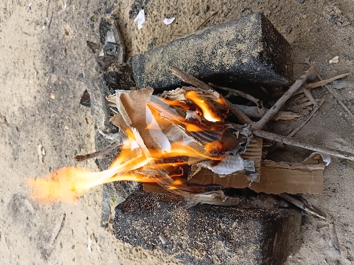 A small fire burns on a sandy surface, contained within makeshift boundaries of two dark, sandy bricks. The fire consumes cardboard and small twigs. Flames rise from the center, predominantly yellow with hints of orange. The surrounding sand is light in color, scattered with debris. A discarded cigarette butt is visible among the sand.
