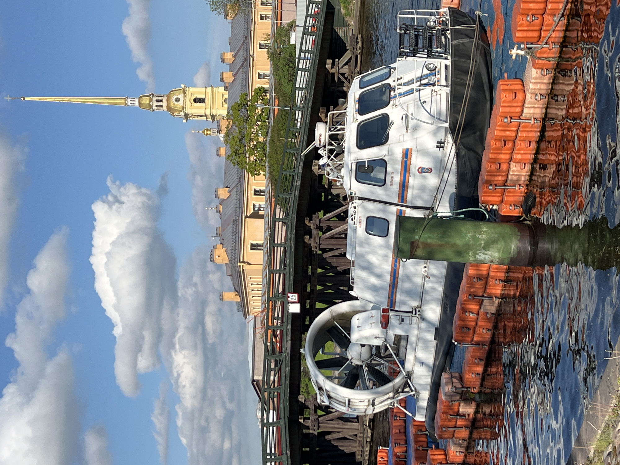 A partially obstructed and rotated outdoor shot shows a white hovercraft docked on a river in Saint Petersburg, Russia, on a partly cloudy day.
The hovercraft, labeled with "TEPTPOXOD 01," is docked against a row of orange floats. Its features include a large fan at one end, a cabin with windows, and a green exhaust. The water reflects the hovercraft and the floats.
Overhead, a pedestrian bridge spans the river. The background reveals a building with a golden spire, indicative of Saint Petersburg's architectural style. The sky is filled with scattered clouds.