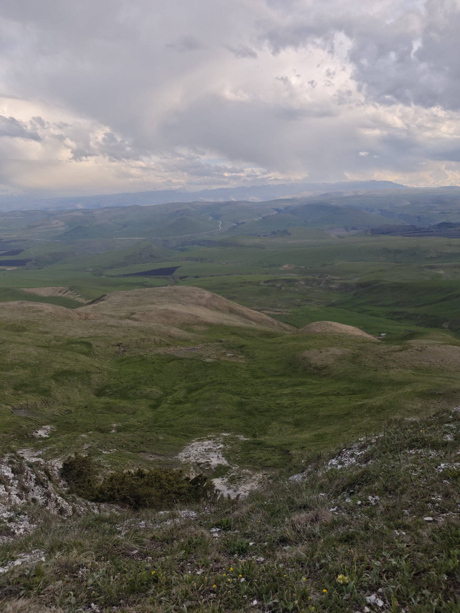 The image depicts a vast, hilly landscape in Kichmalka, Russia, under a cloudy sky. The foreground shows a grassy slope with scattered rocks and small patches of vegetation. Midground consists of rolling hills covered in green grass, with some areas showing patches of brown. In the background, distant mountains are visible under the cloudy sky. A road is faintly visible winding through the landscape. There are no people visible in the image.