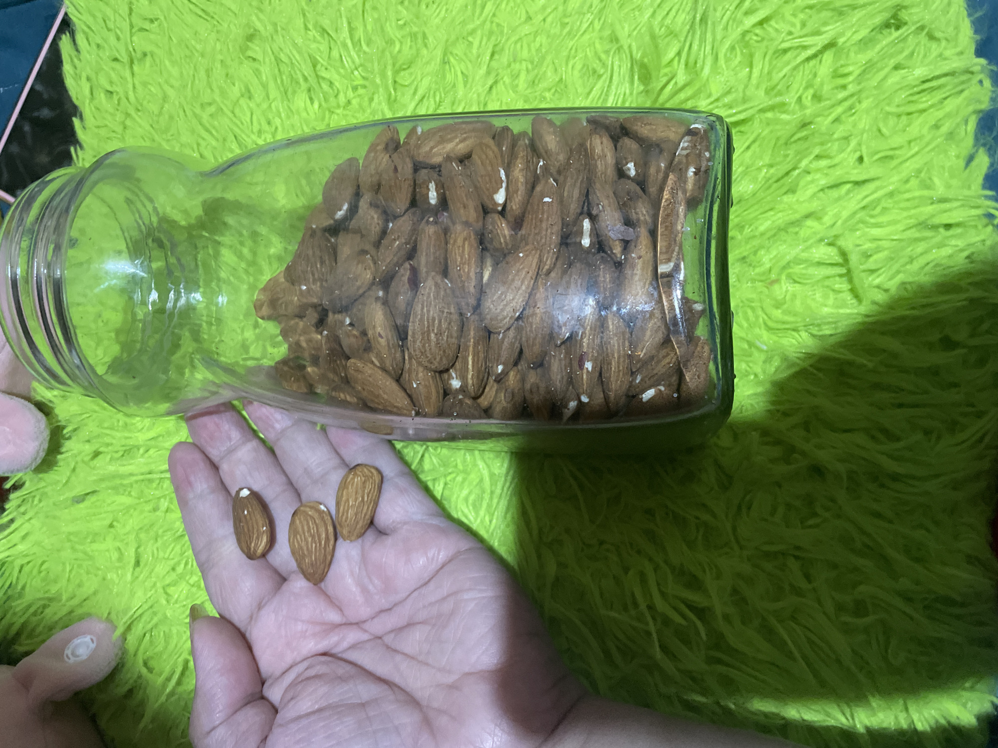 A jar of almonds is on its side on a bright green, shaggy surface. A person's hand is beneath the opening of the jar, holding three almonds. The location is Davao City, Philippines.