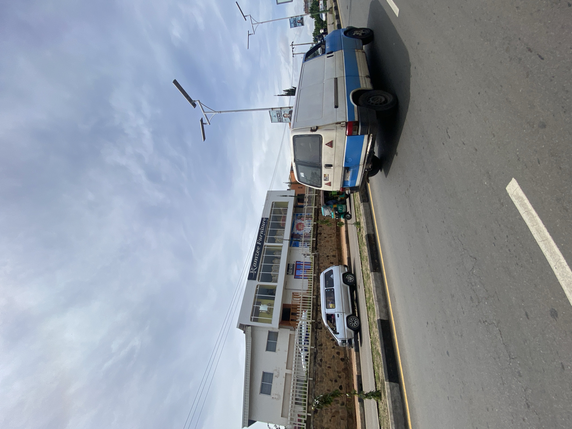 The image shows a street scene in Jos, Nigeria. In the foreground, a grey asphalt road with white and yellow lane markings stretches towards the horizon. On the right side of the road, a blue and white panel van is driving away from the camera. On the left side of the road, a white minivan is parked on the side of the street. Behind the minivan, there is a building with a sign that reads "Ramzee Furniture". The building has large windows and a white facade. Beyond the building, there are trees and a cloudy sky. The environment appears to be an urban setting with buildings and infrastructure common in Nigerian cities. There are no people clearly visible in the scene, though there may be occupants in the vehicles. The overall impression is a snapshot of everyday life on a street in Jos.