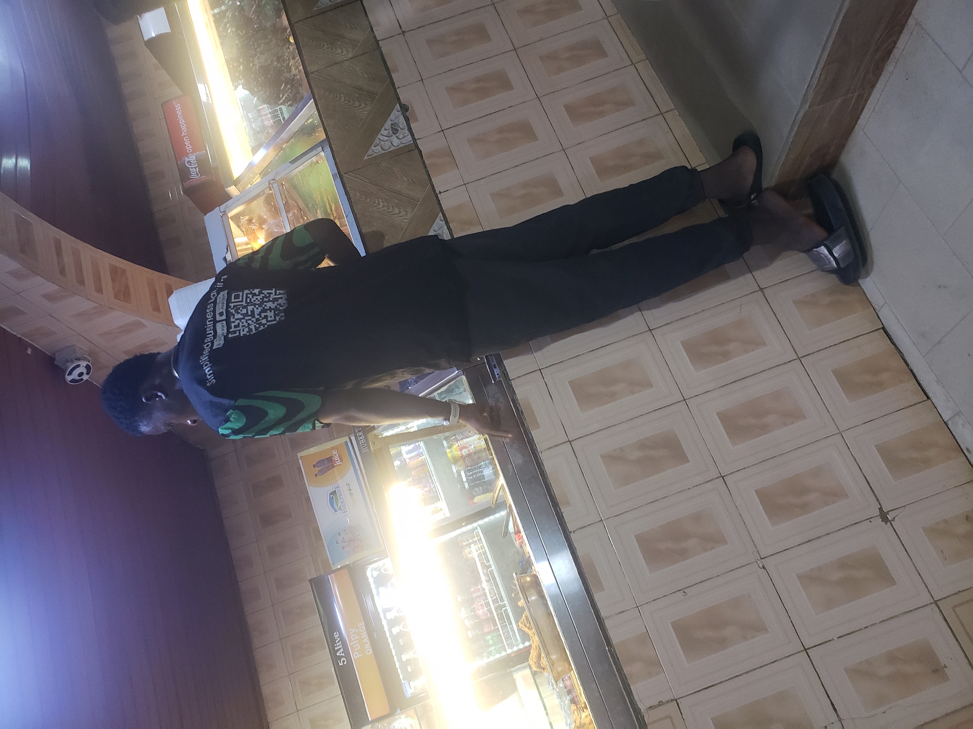 This indoor image captures a person standing beside illuminated commercial display cases, likely within a shop or convenience store in Barden Yana, Nigeria. The individual, a male, is seen from a mid-height perspective, wearing a black short-sleeved top with a green pattern on the right side and sleeve, and dark trousers. His left arm is extended towards the display cases, suggesting interaction or observation of the products.
The person's shirt prominently features white text that reads "Simplified Business" accompanied by a QR code, beneath which faint "Google Play" and "App Store" icons are visible, indicating a potential mobile application or service.
The setting includes two brightly lit display cases, suggesting they contain refrigerated items such as drinks. The intense light from these cases creates some glare. On these cases or the products within, text such as "5 Alive Pulpy ORANGE," "FARM PRIDE juice," and "HUBERT juice" is discernible. A red sign with "Coca-Cola open happiness" is also visible in the background.
The floor is covered with light-toned rectangular tiles, each containing a darker, square-within-a-square pattern. A plain wall lines the right side of the frame, and parts of the ceiling or upper wall structure are visible on the left. The prominent artificial lighting implies the photo was taken indoors where natural light is limited, possibly in the evening or at night. The overall atmosphere points to a casual, local commercial establishment. The image itself is rotated 90 degrees counter-clockwise.