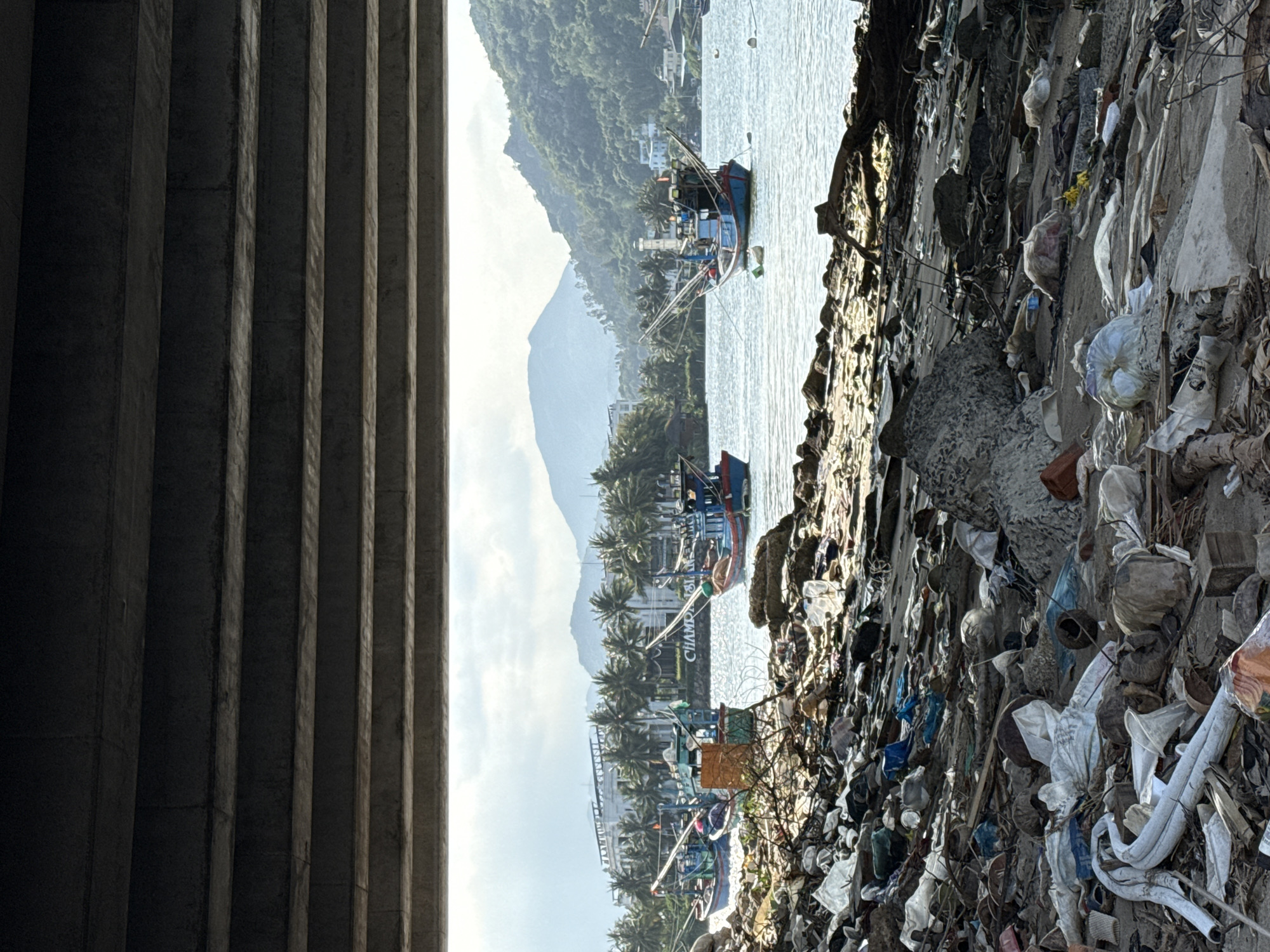 The image depicts a scene of significant pollution along the coastline of Nha Trang City, Vietnam, specifically in Bac Nha Trang Ward. The foreground is dominated by a large accumulation of trash, including plastic bags, bottles, debris, and what appear to be discarded fishing nets and ropes, strewn across the sandy shore. This waste creates an unsightly and environmentally damaged landscape.
Beyond the polluted foreshore, a body of water is visible, likely the Nha Trang Bay. Several fishing boats, characterized by their distinctive blue and red hulls and prominent fishing gear, are anchored or navigating on the water. In the background, a lush green hillside with dense vegetation rises, dotted with buildings and palm trees, suggesting a coastal urban or semi-urban environment. The presence of a large concrete structure, possibly a bridge or overpass, frames the upper portion of the image, casting a shadow over the scene and adding a sense of scale. The sky is partly cloudy, with the sun casting light and shadows across the water and land. The overall impression is one of natural beauty marred by severe human-induced pollution.