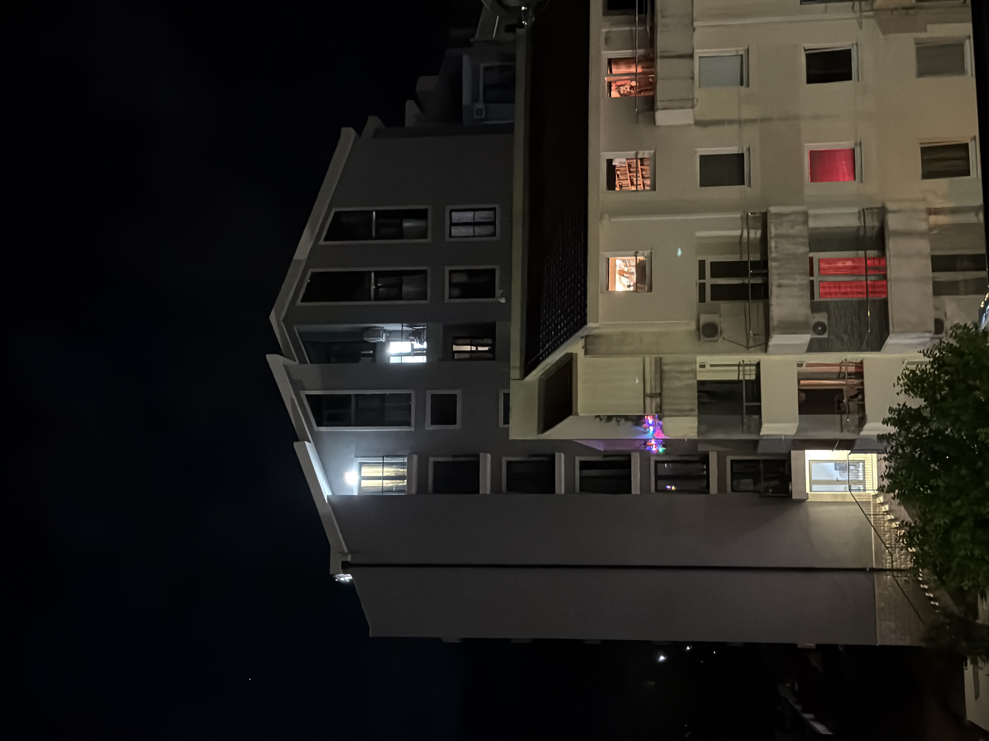 The photograph shows two buildings in Budva, Montenegro at night. The building on the left is gray with a distinctive angled roofline and multiple windows, some illuminated. The building on the right is taller and white, with numerous windows, some showing interior light, including one with red curtains. Colorful lights are also visible on one of the floors. To the right, foliage from a tree is visible. The sky is dark.
