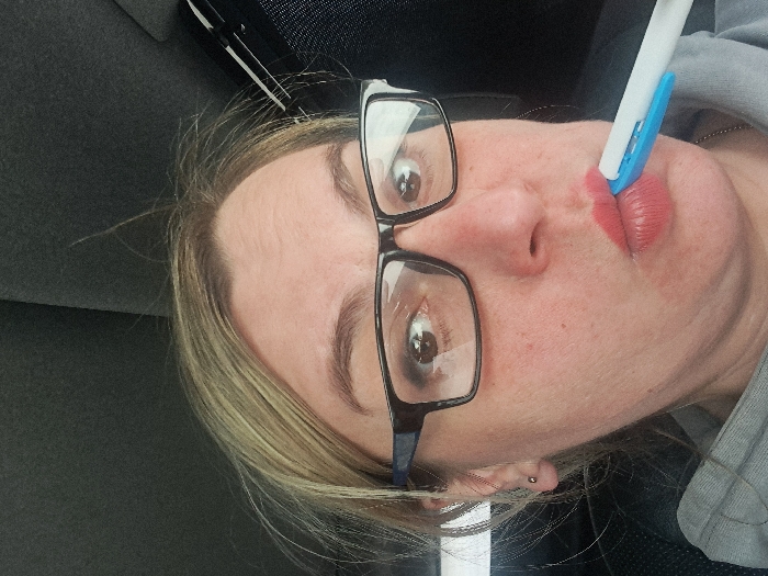 An adult female is depicted in a close-up, frontal portrait, situated within what appears to be the interior of a vehicle in Novaya Derevnya, Russia. She has blonde hair, wears black-framed eyeglasses, and a small, spherical stud earring is visible in her left earlobe. Her eyes are wide open, directed towards the camera, with visible light reflections in the pupils. Her lips are pursed and coated with reddish lipstick, actively gripping a white pen with a blue segment horizontally between them. She is dressed in a grey hooded garment, partially visible. The background is a dark, textured fabric consistent with a car's headliner, with a dark structural element visible in the upper right.