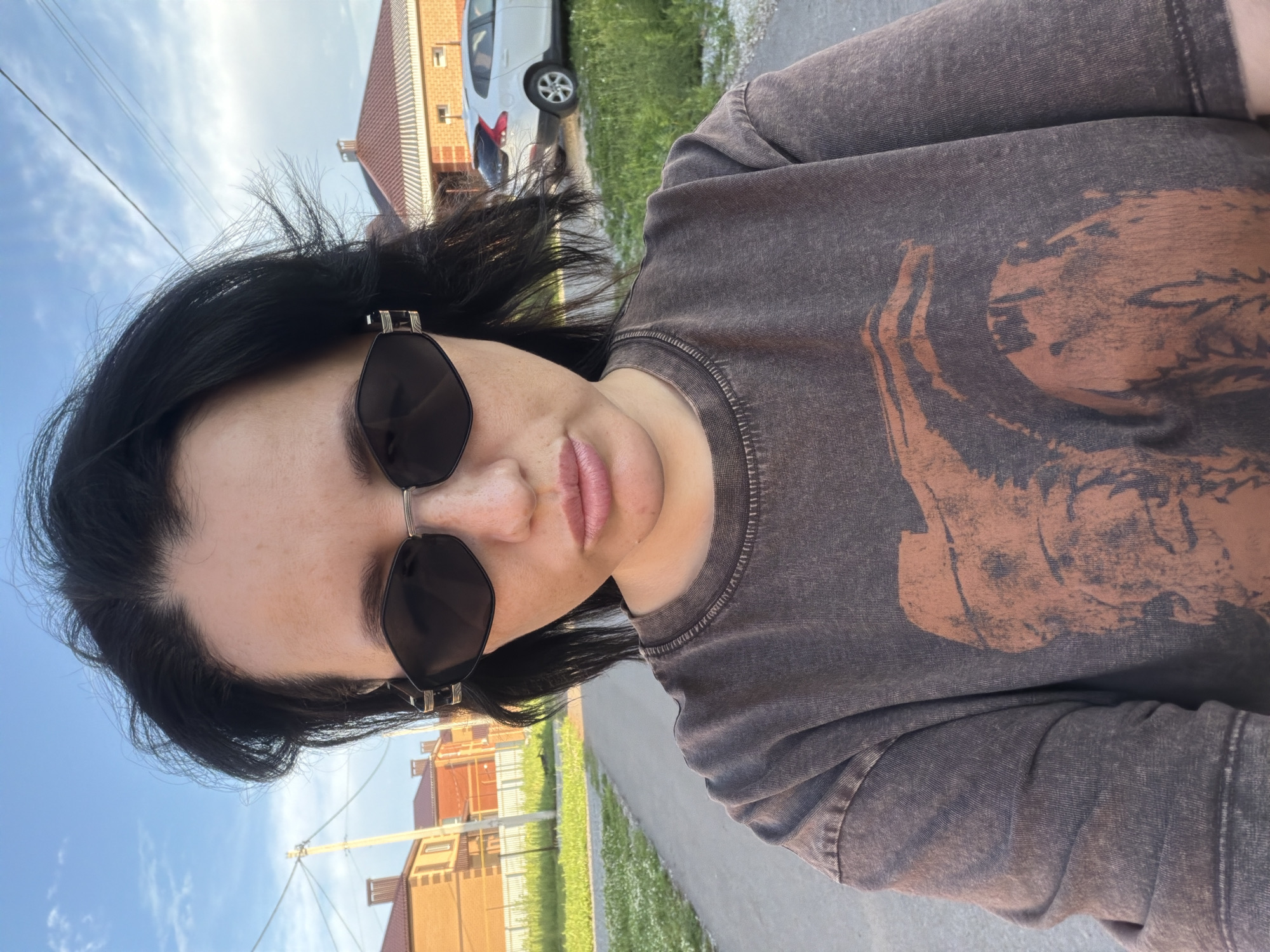 In Шмидтово, Russia, a woman with black hair and sunglasses is taking a selfie on a paved road. She is wearing a dark brown t-shirt with a faded graphic print of an elephant. The background shows houses with red-tiled roofs, green grass, and a blue sky with some clouds. A white car is parked on the side of the road.