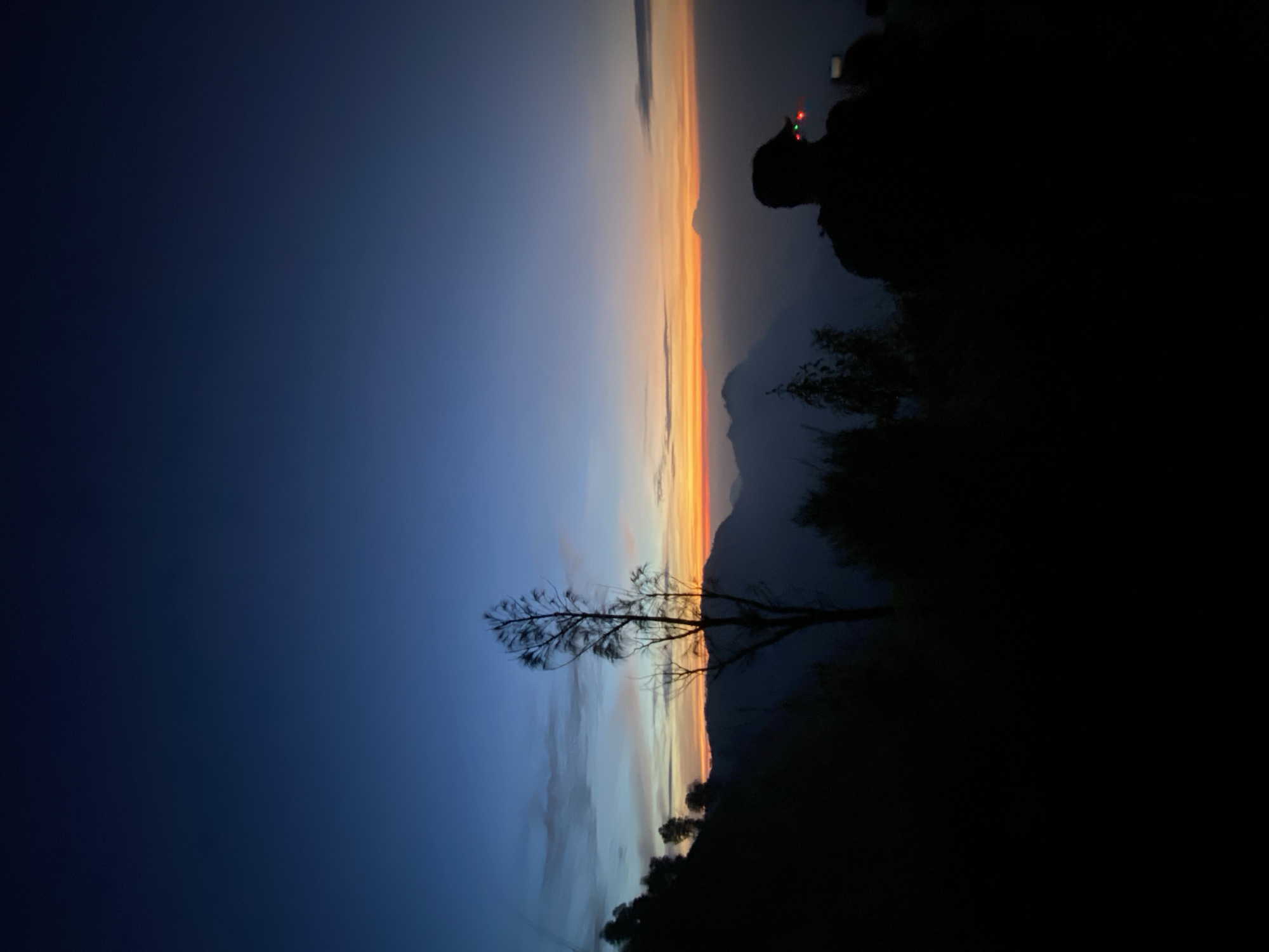 Captured at dusk in Cemorolawang, Indonesia, the landscape features a stark contrast between the darkening blue sky and the vibrant horizon. A silhouetted tree stands prominently, with mountains and vegetation fading into shadow. A person, possibly wearing a headlamp, is visible on a ridge.