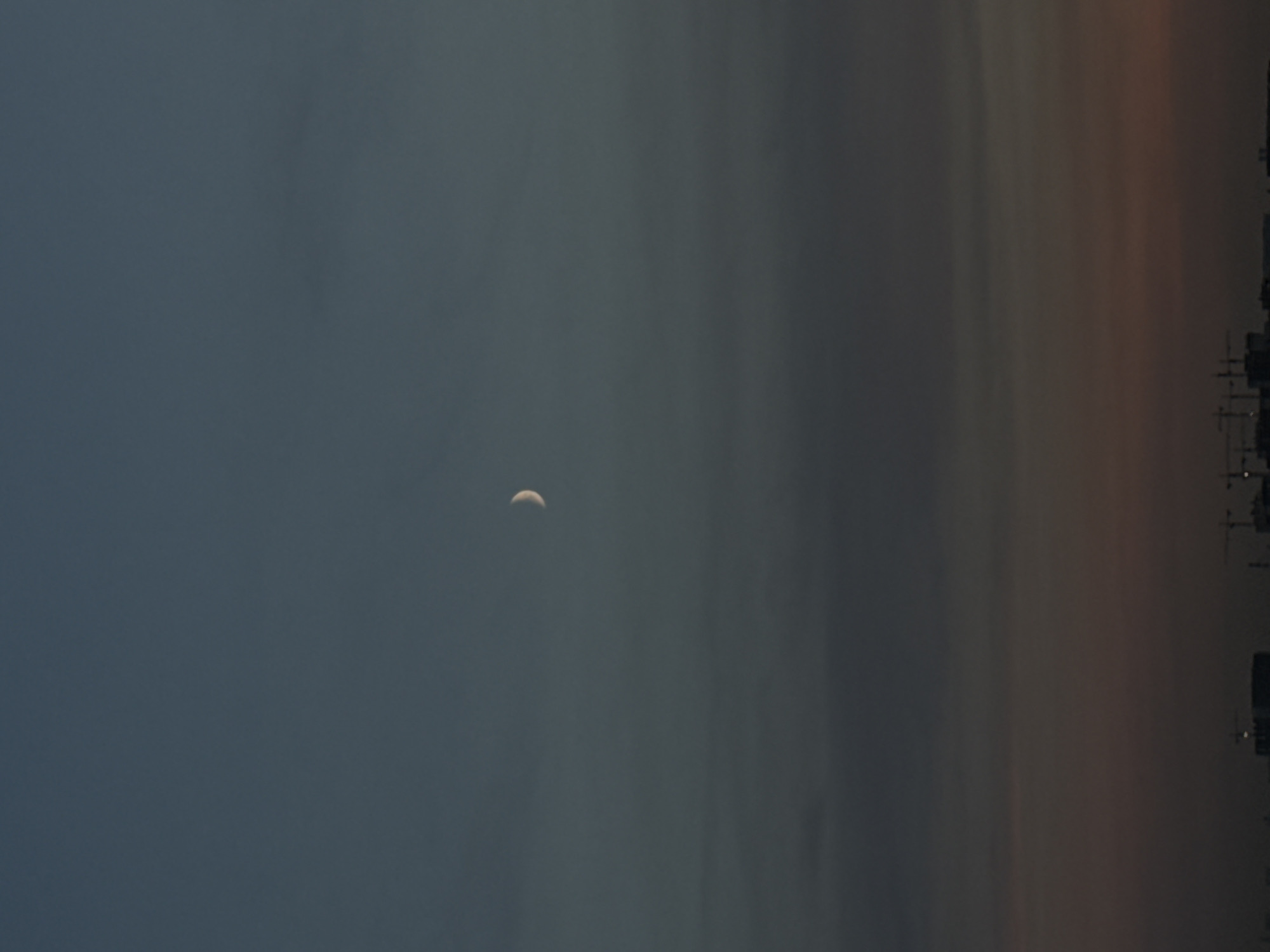This is a vertical, eye-level, medium shot photograph taken in Lyubertsy, Russia. The image depicts a hazy, dusky sky with a crescent moon visible in the upper middle portion of the frame. The lower third of the image shows the silhouette of buildings and construction cranes against a gradient of muted colors, transitioning from dark grey to a faint reddish-orange hue near the horizon. The overall atmosphere is calm and still, with no people or discernible activity present. The sky is predominantly a deep, muted blue-grey, with subtle cloud formations contributing to the soft, diffused light. The urban landscape below is rendered in dark, indistinct shapes, emphasizing the vastness of the sky and the quietude of the evening.