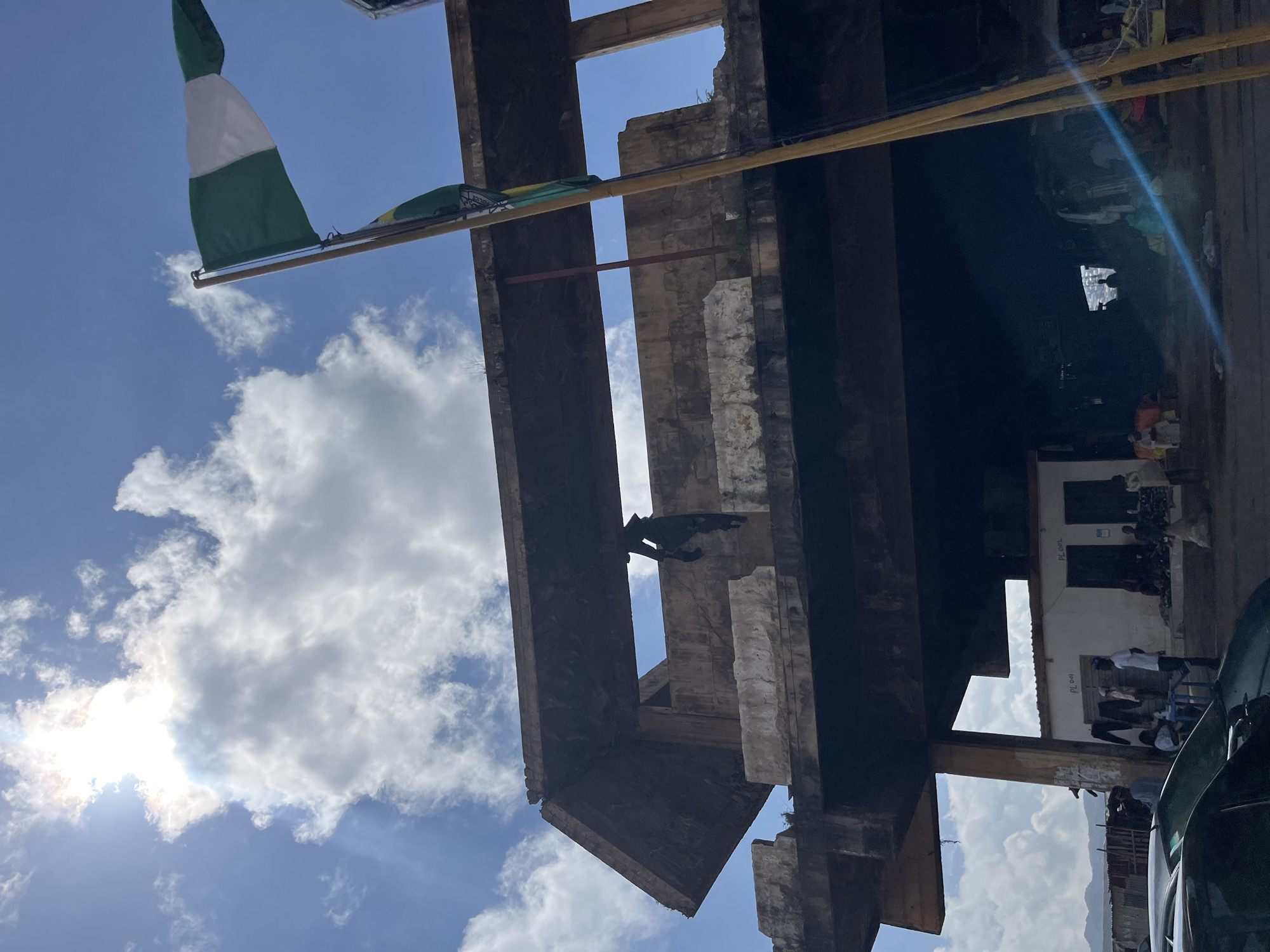 In Jos, Nigeria, under a bright, cloudy sky, the image captures an unfinished concrete structure. A Nigerian flag waves proudly from a wooden pole in the foreground, with another smaller flag below it. A person in dark clothing is visible hanging from the upper level of the concrete building, partially obscured by shadows. The lower level of the structure houses a small marketplace with several people, stalls displaying clothing, and parked cars. The overall scene depicts a blend of construction, commerce, and human activity in an urban setting.