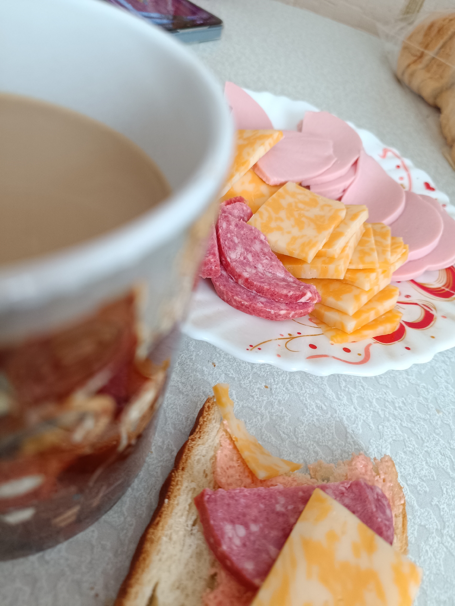 The image shows a breakfast scene on a table. A partially visible cup of coffee is on the left. Next to it is a white plate with sliced meat and cheese. A half-eaten sandwich with bread, spread, sausage, and cheese is in the foreground. A phone sits on the table and a loaf of bread is in the upper right. The location is implied to be in Tobolsk, Russia, but this cannot be directly ascertained from the visual elements of the image.