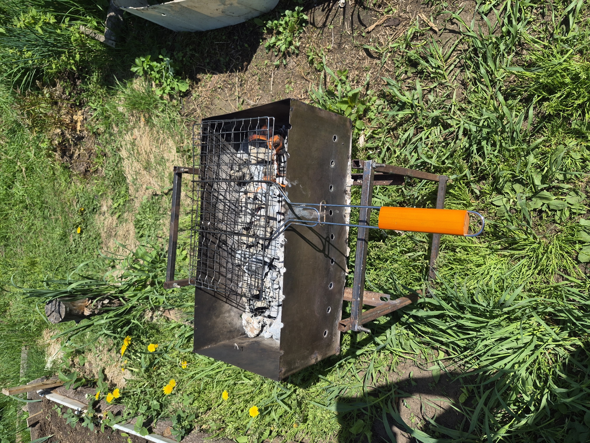 The image shows a rectangular charcoal grill, holding a wire grill grate with an orange handle. The grill contains ashes and a few embers. It is positioned on green grass, possibly in a yard or garden setting. Other plants are visible in the background. The location appears to be outdoors in a natural environment.