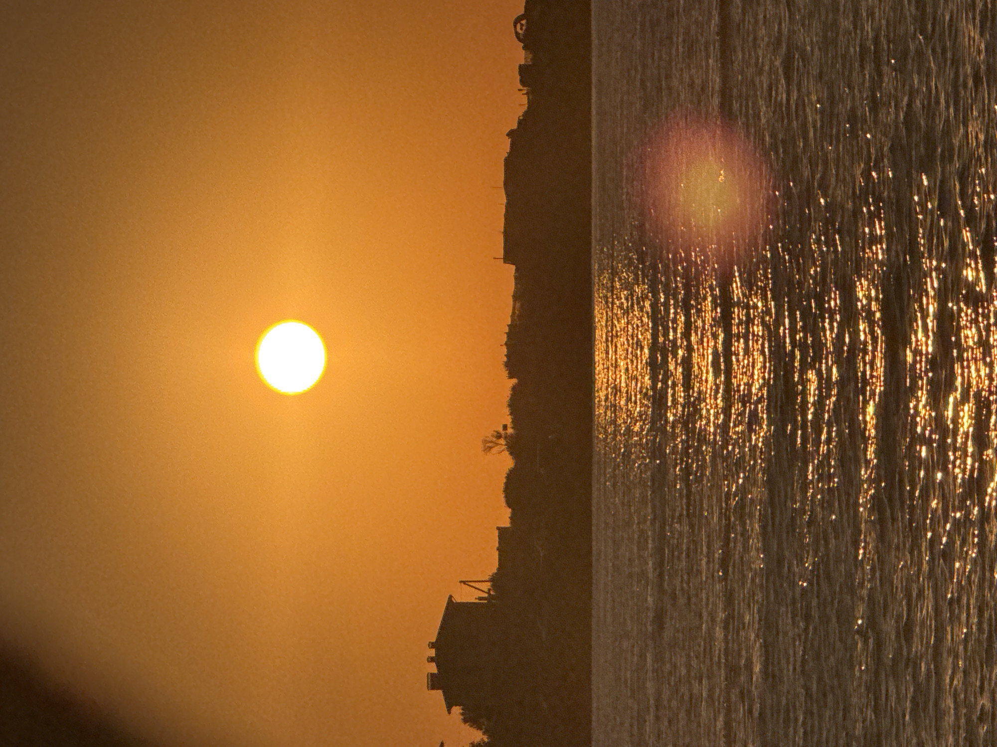 An evening shot captures a wide body of water, likely the Black Sea, in front of a silhouetted city skyline. The sun, a bright orange orb, is low in the sky, casting a warm, orange glow across the scene. Its reflection shimmers on the rippling surface of the water, creating a path of light that extends towards the viewer. The city in the background is a dark, indistinct silhouette against the vibrant sky. Buildings, some with peaked roofs, are visible, along with trees and other structures, suggesting an urban or coastal settlement. The overall impression is one of tranquility and natural beauty, with the setting sun dominating the frame. The location is Sevastopol, Ukraine, a port city known for its beautiful coastal scenery and historic significance.