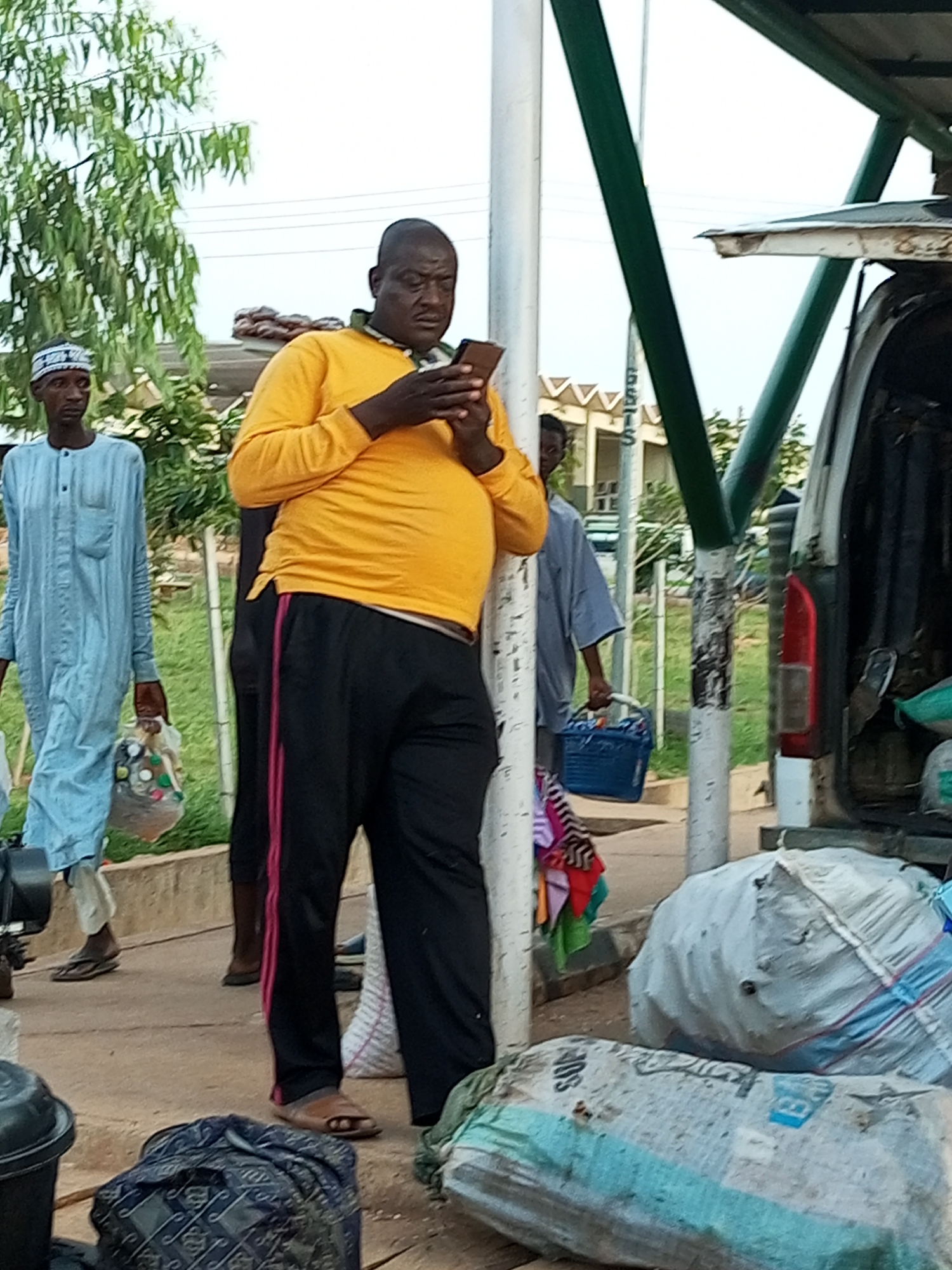 The image depicts an outdoor scene in Gombe, Nigeria, likely near a transportation hub or market area, as evidenced by the presence of a vehicle and large bags of goods. In the foreground, a man wearing a bright yellow long-sleeved shirt and black pants with pink stripes is engrossed in his mobile phone. He is standing next to a white pole. To his left, another man, wearing a light blue traditional garment and a white skullcap, holds a plastic bag filled with items. Further in the background, a partially obscured person in a grey shirt is visible, and to the right, the back of a white van is open, revealing various bags and items. Large, wrapped bales or bags of goods are also prominent in the foreground and midground. The environment suggests an active area with commercial activity.