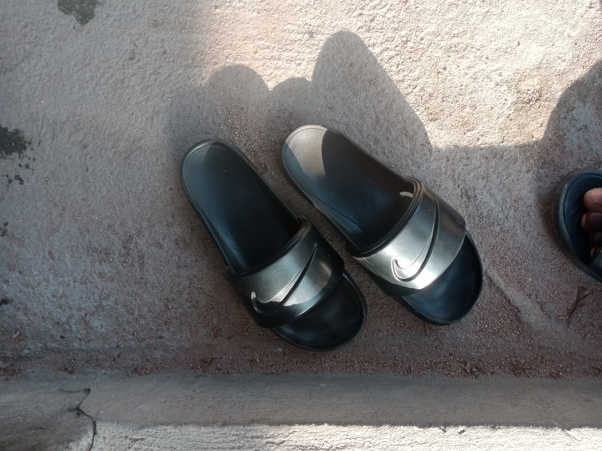 A pair of black Nike slide sandals are placed on a rough, sandy, outdoor surface. The sandals are positioned side-by-side, with the left sandal slightly closer to the bottom edge of the frame. Both sandals feature a metallic silver Nike swoosh logo across the straps. Shadows cast from an unseen object fall across the sandals and the ground. A small portion of a person's foot, wearing a black sandal, is visible on the right side of the frame. The ground appears to be a mixture of sand and small pebbles, with some darker patches that might be water stains or debris. The environment suggests an outdoor location, possibly a walkway or courtyard. The lighting indicates it is daytime, with strong sunlight creating distinct shadows. The scene appears to be located in Jalingo, Nigeria, though no specific landmarks are visible to confirm this definitively.