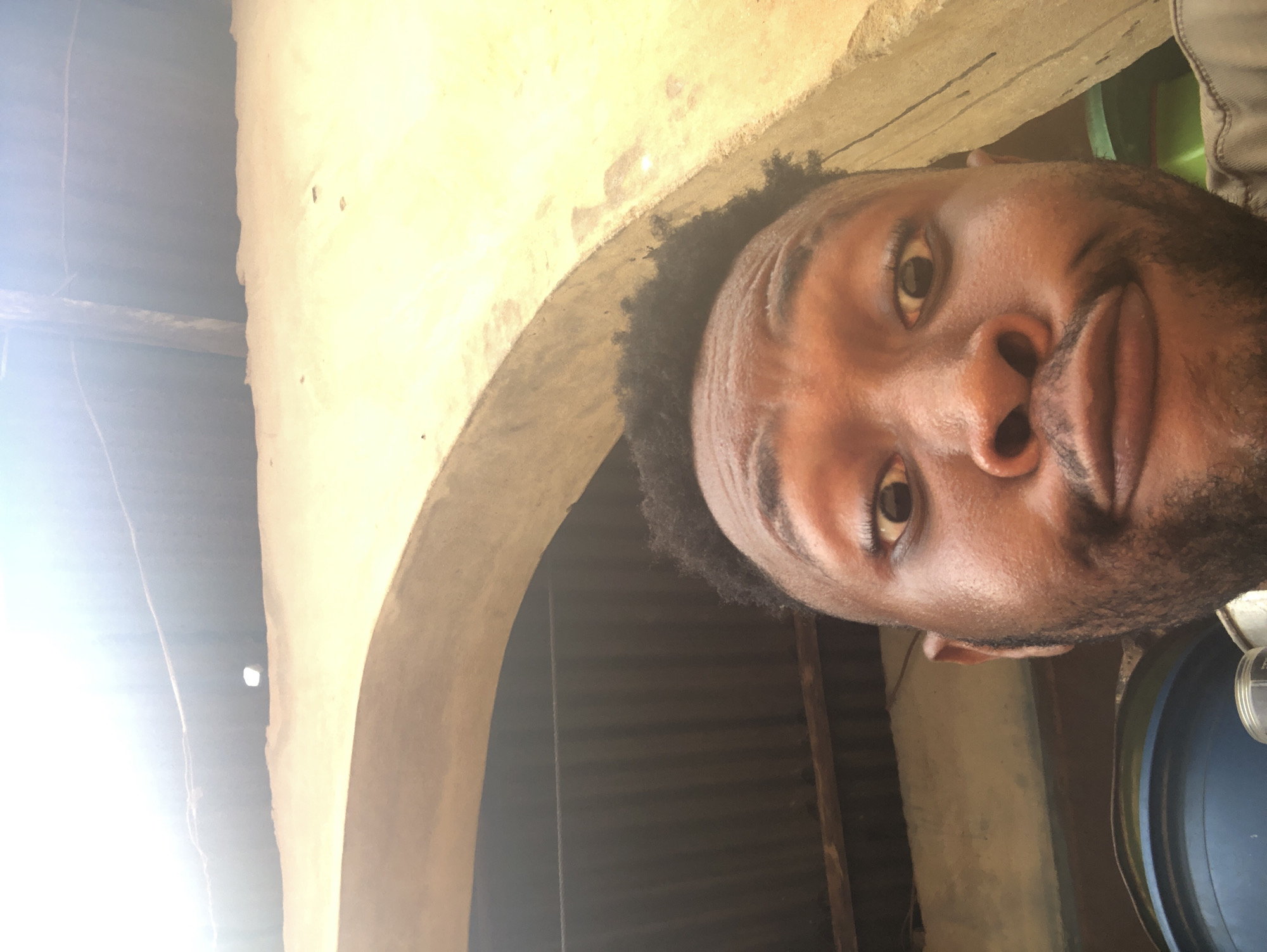 A close-up shot shows a Black man's face, tilted toward the camera. The background includes a rough, yellow-toned wall and what appears to be an arched doorway or opening. Behind the opening, there are dark, corrugated metal sheets. To the right of the man's head, a portion of a blue plastic container is visible. The location is Unknown, Unknown.