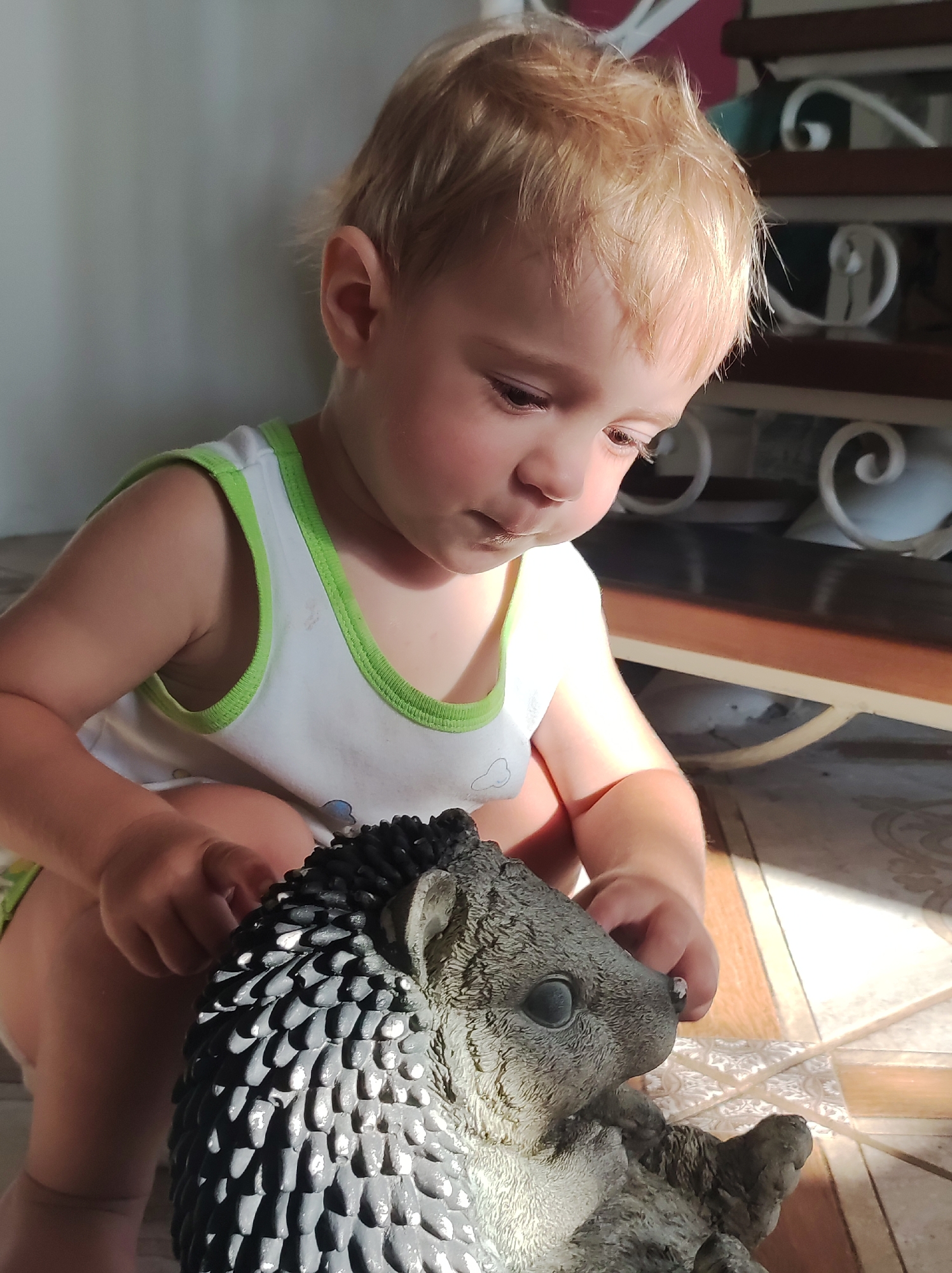 A fair-skinned toddler with blonde hair is crouched down, looking at a small, grey, spiky hedgehog figurine. The toddler is wearing a white tank top with a green trim. Their right hand is touching the figurine's nose. Sunlight streams from the right, illuminating the toddler's face and the figurine. The background shows a tiled floor and what appears to be a wooden shelf or staircase with decorative metalwork.
