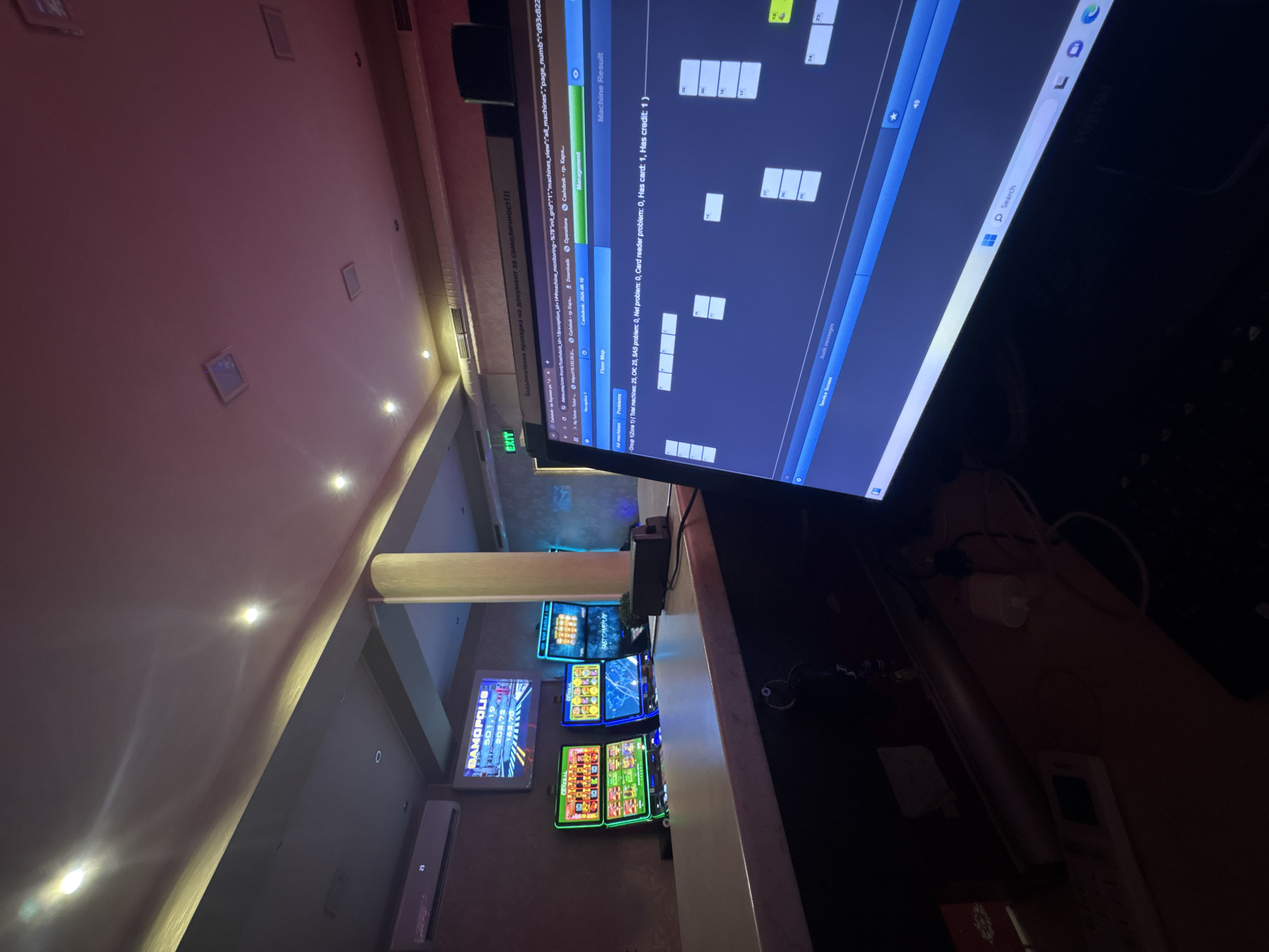 The image depicts the interior of a casino or gaming establishment, likely located in Karlovo, Bulgaria. In the foreground, a computer monitor displays a software interface, possibly for managing the casino's machines. The screen shows a "Floor Map" with various machine statuses indicated by colored rectangles. A web browser is also visible, with URLs suggesting it's related to machine monitoring and cash desk operations.
In the middle ground, a counter separates the computer area from the gaming floor. Behind the counter, several slot machines are visible, some active and displaying game graphics. One prominent display screen above the slot machines shows "DAMOPOLIS" with a series of numbers. The environment appears to be dimly lit, with overhead lighting and accent lighting creating a specific ambiance.
In the background, an air conditioning unit is mounted on the wall, and an "EXIT" sign is illuminated. There are also pillars and ceiling details suggestive of a commercial space. The overall scene conveys a sense of a functional gaming environment, with the computer system likely being used for operational oversight. There are no people visible in the image.