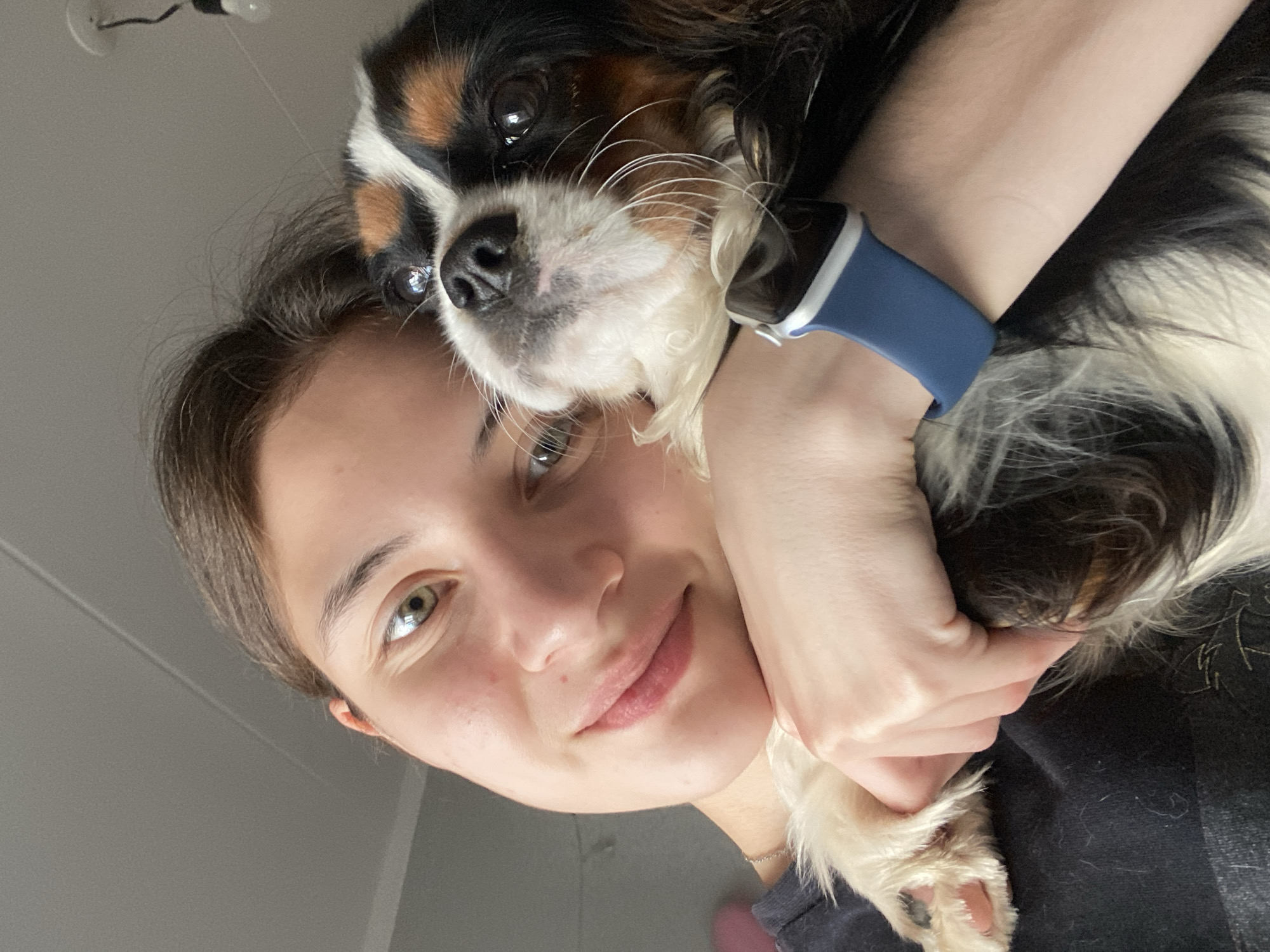 In a close-up, indoor photograph taken from a high angle, a young Caucasian female with brown hair is holding a small Cavalier King Charles Spaniel. The woman is wearing a dark t-shirt and a silver necklace. She has light skin, green eyes, and is smiling softly. The dog, predominantly white with black and tan markings, is nestled against her chest. Its muzzle and nose are visible, and its ears are characteristic of the breed. The woman's left arm is wrapped around the dog, and her right hand is holding its front paw. A blue smartwatch is visible on her left wrist. The background is blurred, showing a neutral-colored ceiling with a light fixture and some indistinct objects. There are no explicit indicators of the location being Votkinsk, Russia.