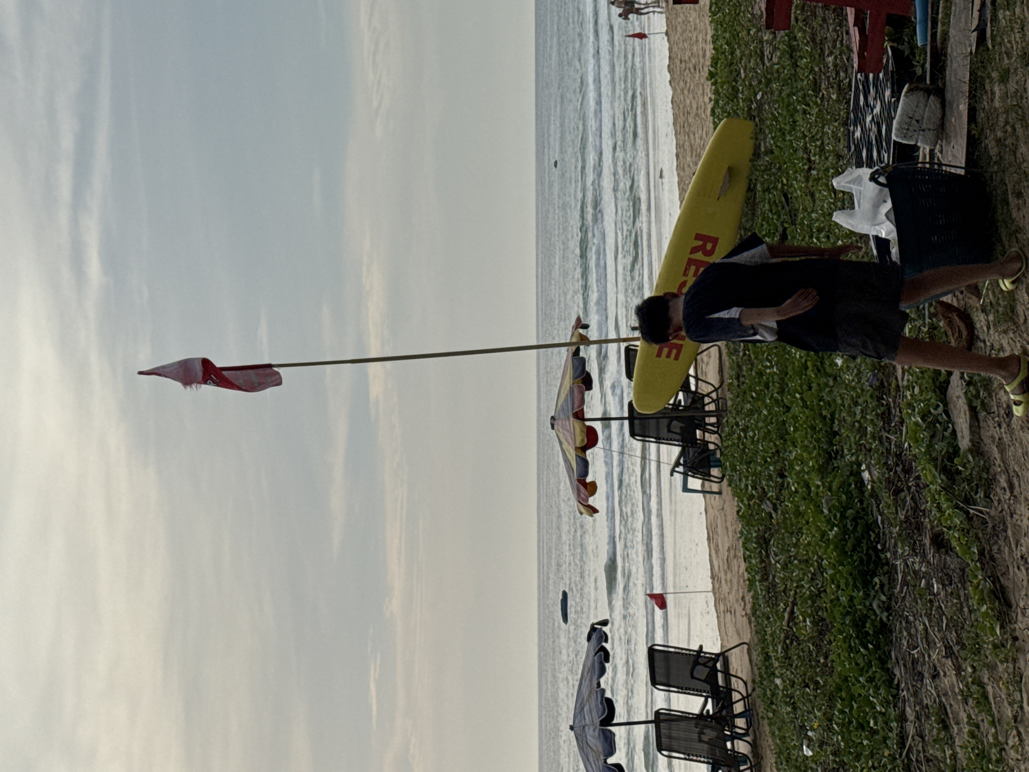 This image captures a scene on Karon Beach, Thailand, on an overcast day. In the foreground, a young male wearing dark shorts and a blue and white shirt stands facing away from the camera. He appears to be walking towards a black basket filled with white plastic bags. To his left, a yellow surfboard with the word "RESCUE" printed in red is leaning on the sand. Behind the surfboard, several beach chairs and umbrellas are arranged on the sand near the water's edge. A small red flag is planted in the sand between two chairs. The ocean is visible in the background with gentle waves. In the distance, a thin flag on a pole flies in the sky. The overall environment is a beach setting with vegetation bordering the sandy area.