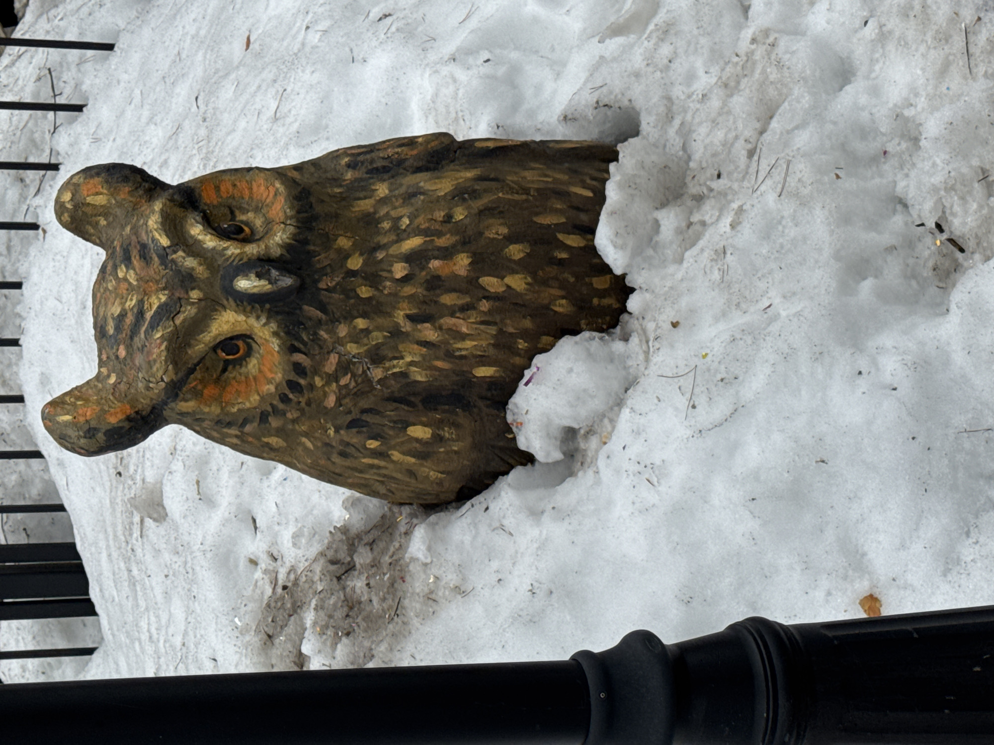 A carved wooden owl head emerges from a snowdrift in Chaikovsky, Russia. The owl is painted in brown and gold with orange accents around the eyes. It appears to be near a black metal fence and a black metal post. The scene suggests a park or garden setting covered in snow.