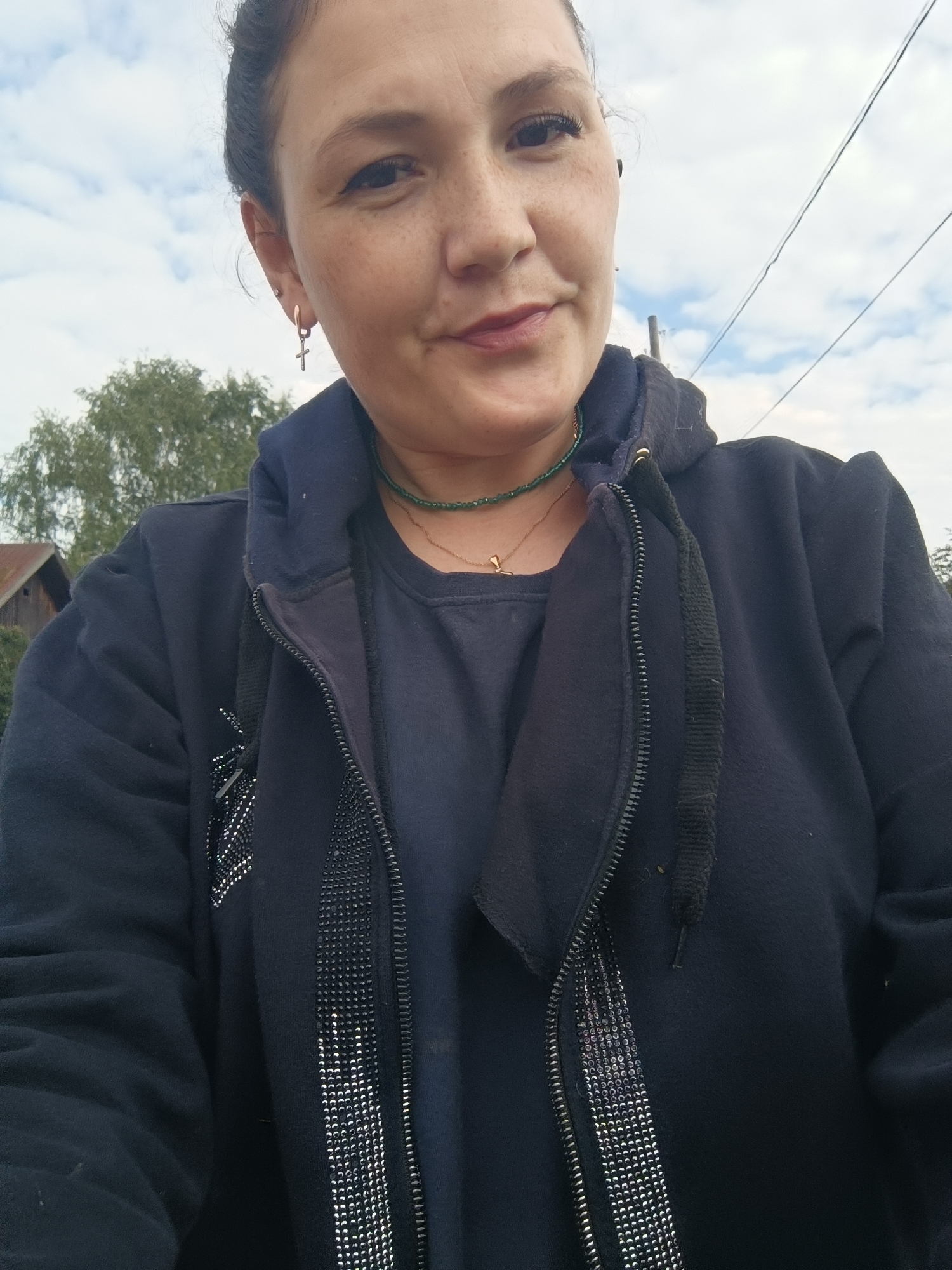 A woman with dark hair pulled back is pictured from the chest up, looking slightly off-camera. She has freckles across her nose and cheeks and is wearing a dark blue hooded jacket over a dark blue t-shirt. A thin green beaded necklace with a small gold pendant is visible around her neck, and she has a gold hoop earring with a cross charm in her left ear. The background consists of a cloudy sky, power lines, and trees with a hint of a building visible on the left. The setting does not appear to be the city of Karsha, Russia.