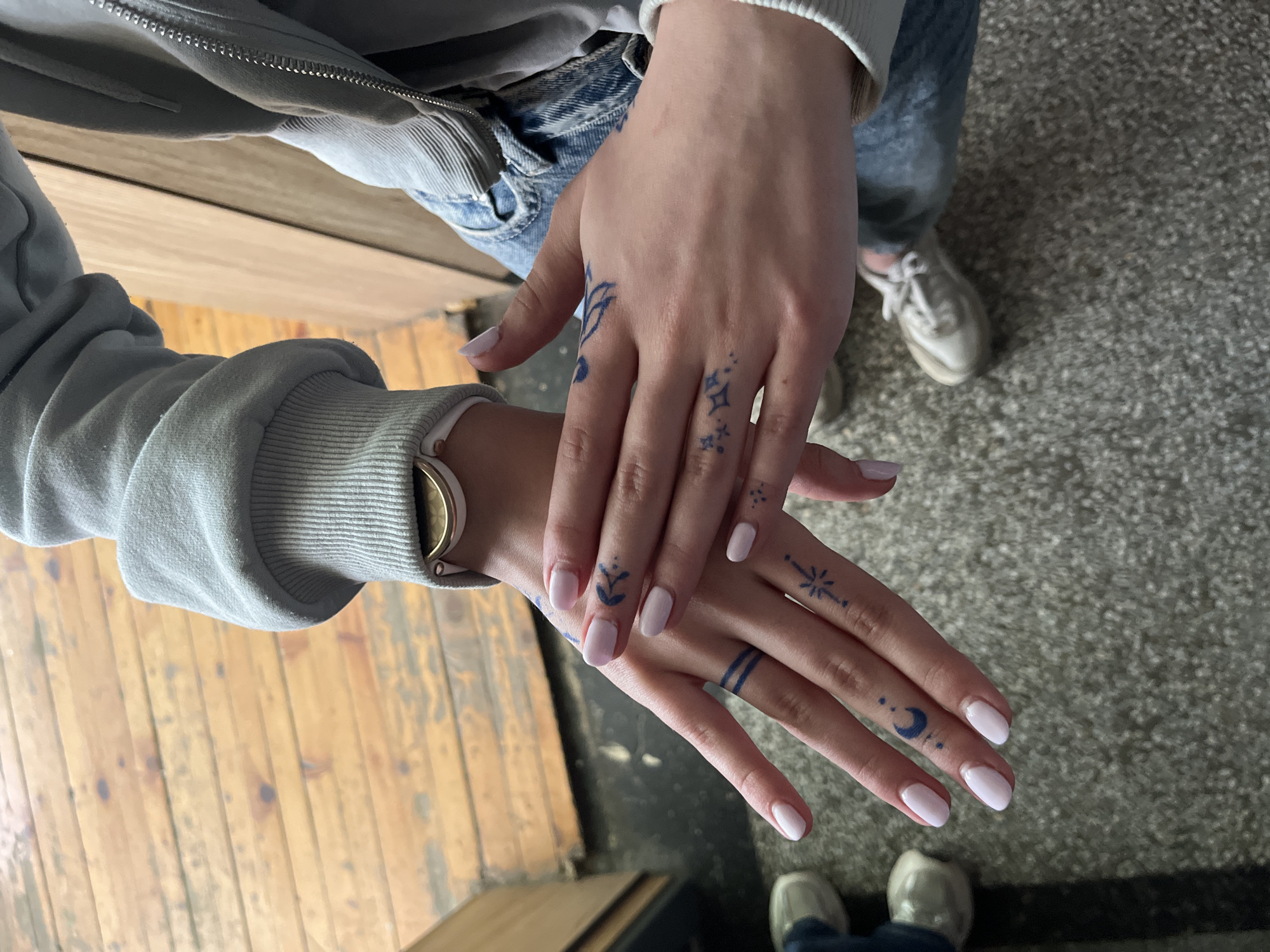 The image displays two hands with light pink painted nails. Both hands are adorned with blue ink tattoos. The left hand has tattoos including a crescent moon with dots and a small leaf design. The right hand features star and diamond designs. The person is wearing a light blue zip-up hoodie and blue jeans. In the background, there's a worn wooden floor and a concrete floor. Two pairs of sneakers are also visible. The specific location within the city of Ruse, Bulgaria, cannot be determined from the visual information provided.