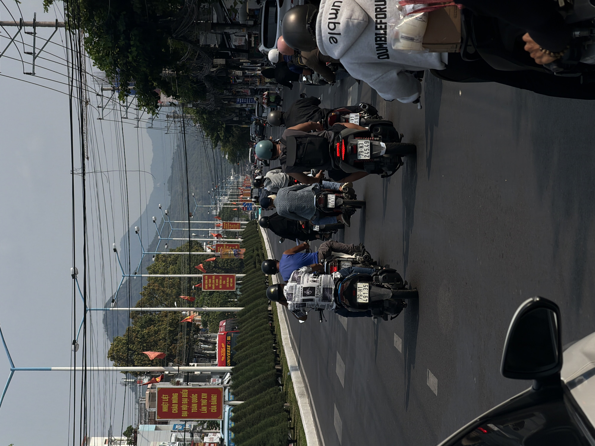 The image captures a busy street scene in Bac Nha Trang Ward, Vietnam, with a convoy of motorcycles moving along a multi-lane road. In the background, mountains are visible under a slightly overcast sky. Along the roadside, there are street lamps and Vietnamese flags attached to poles, and green shrubbery separates the road from the sidewalk. Numerous signs with Vietnamese text are prominently displayed, including political slogans and advertisements. People are riding the motorcycles, wearing helmets and casual clothing. In the foreground, the side mirror and part of a car are visible. The license plates of several motorcycles are clearly visible, and the text on one individual's hoodie reads "Dumbleforu." Overhead, a dense network of power lines spans across the sky.