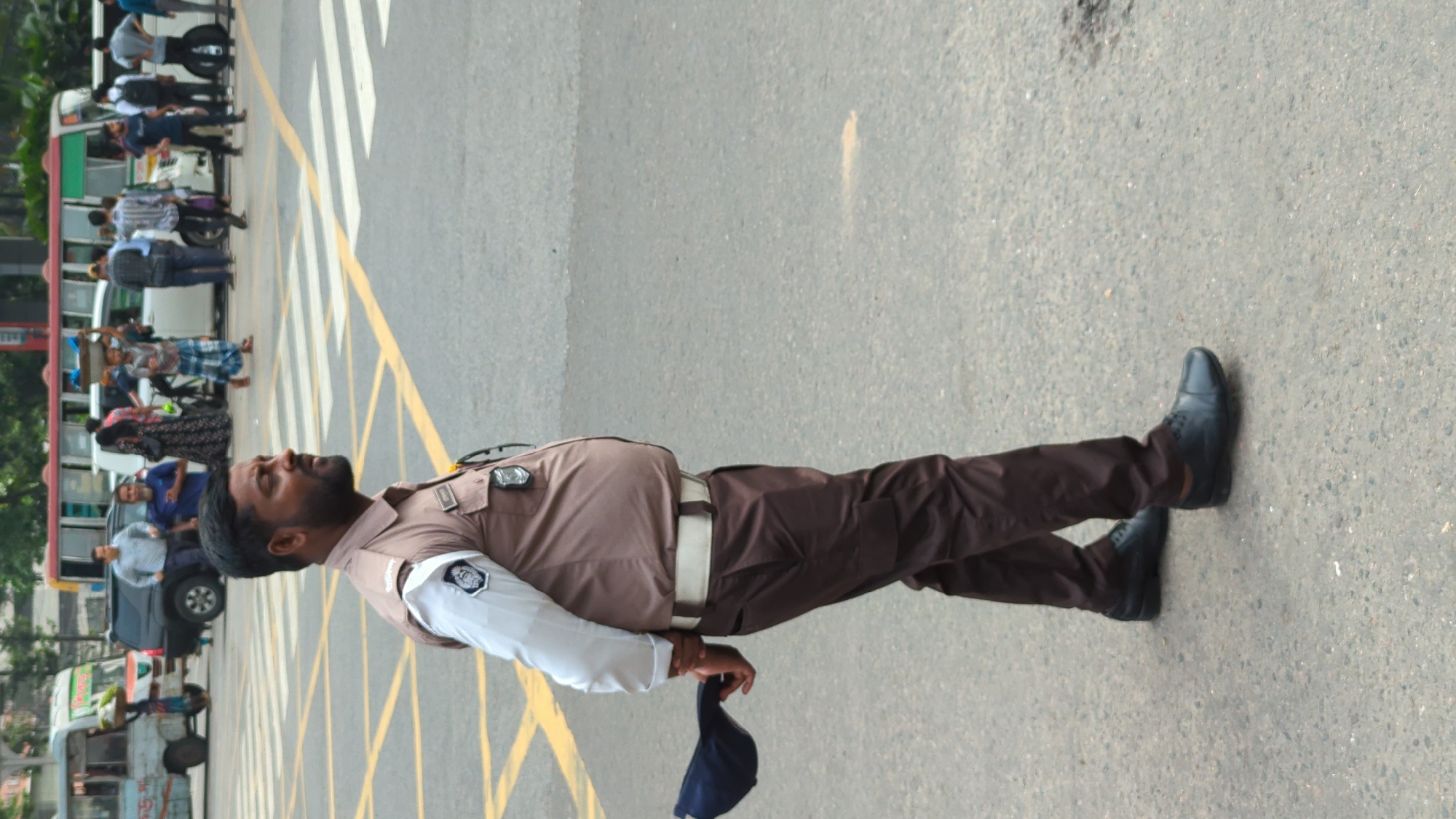 A male traffic officer stands in the middle of a street at an intersection in Dhaka, Bangladesh. He wears a light brown uniform, a white belt, and black shoes, and he holds a dark blue cap in his left hand. The intersection has yellow and white markings. Several buses and people are visible in the background, suggesting a busy urban scene. The environment is likely a central area within the city due to the presence of traffic management and the density of people and vehicles.