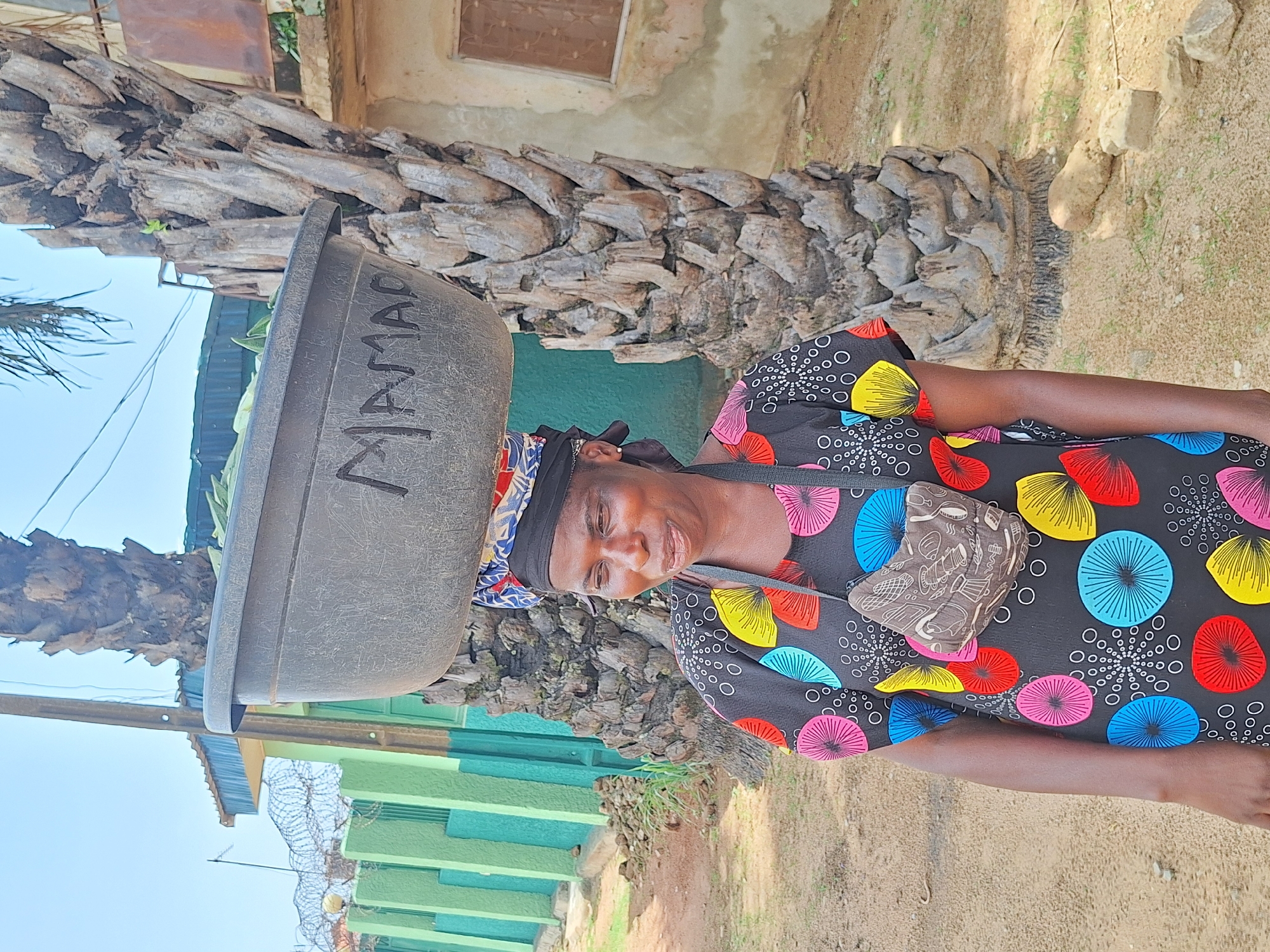A woman carries a large, grey tub marked with the word "MAMA" on her head in Sabon Tasha, Nigeria. She wears a colorful, patterned dress, a black head covering, and a small patterned purse is slung across her body. The background shows a building with green trim, topped with barbed wire, and a palm tree trunk. The ground appears to be sandy soil.