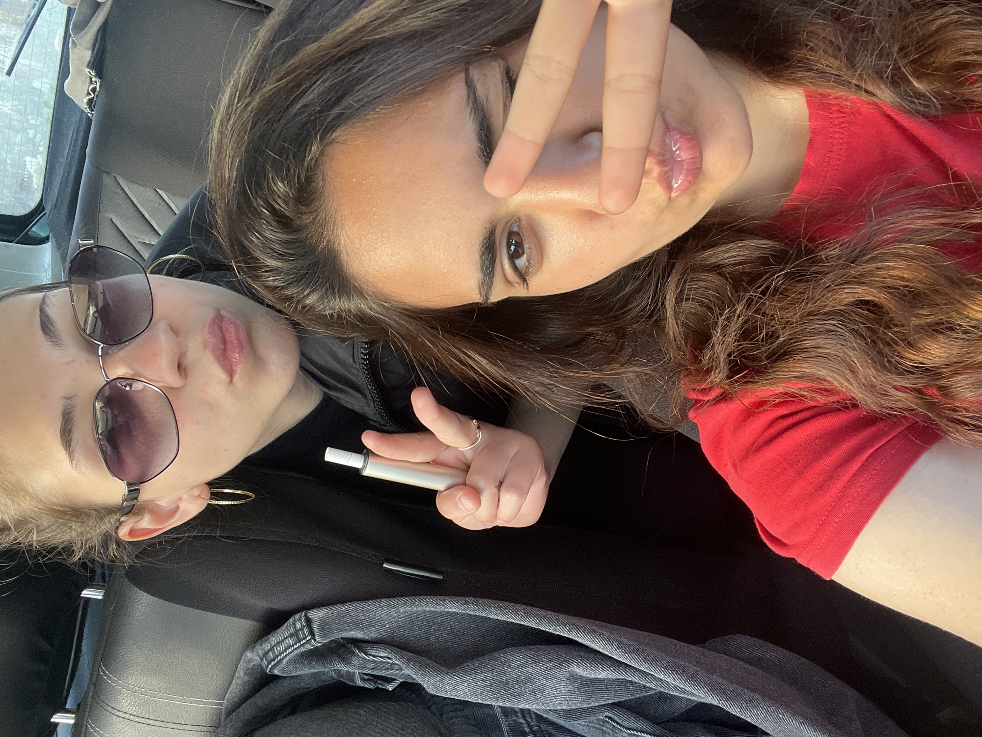 The image shows two young women indoors, likely in a vehicle. One woman is wearing sunglasses, hoop earrings, a black top, and is holding a white electronic cigarette device. The other woman, wearing a red top, is holding up two fingers in a peace sign near her eye. The environment appears to be a car interior with grey leather seats and a denim jacket resting on the seat. The location is Unknown, Unknown.