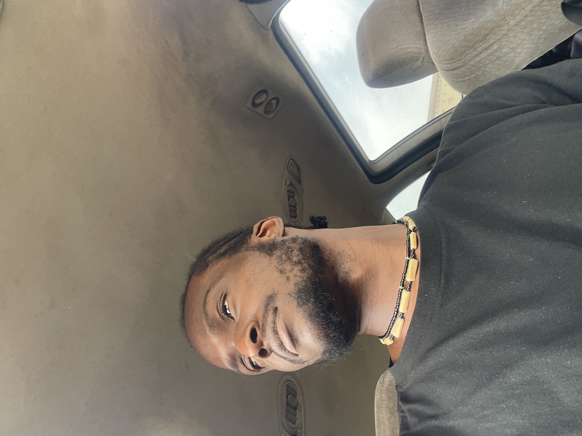 This media file features a close-up, low-angle, portrait-style shot of a young Black man inside a vehicle. The man has braided hair and a beard. He is wearing a black t-shirt and a beaded necklace with light-colored cylindrical beads. He is looking towards the camera with a slight smile.
The interior of the vehicle is visible, with the ceiling panel and a portion of a seat. A sunroof is open, revealing a cloudy sky, suggesting it is daytime, possibly overcast. The overall lighting is soft. The image has a slightly grainy texture.
Based on the provided context, the location is Jos, Nigeria. No visible text is present in the image. The activity appears to be a casual moment, possibly a selfie taken while traveling or waiting in the vehicle. The man's expression conveys a relaxed and approachable demeanor.