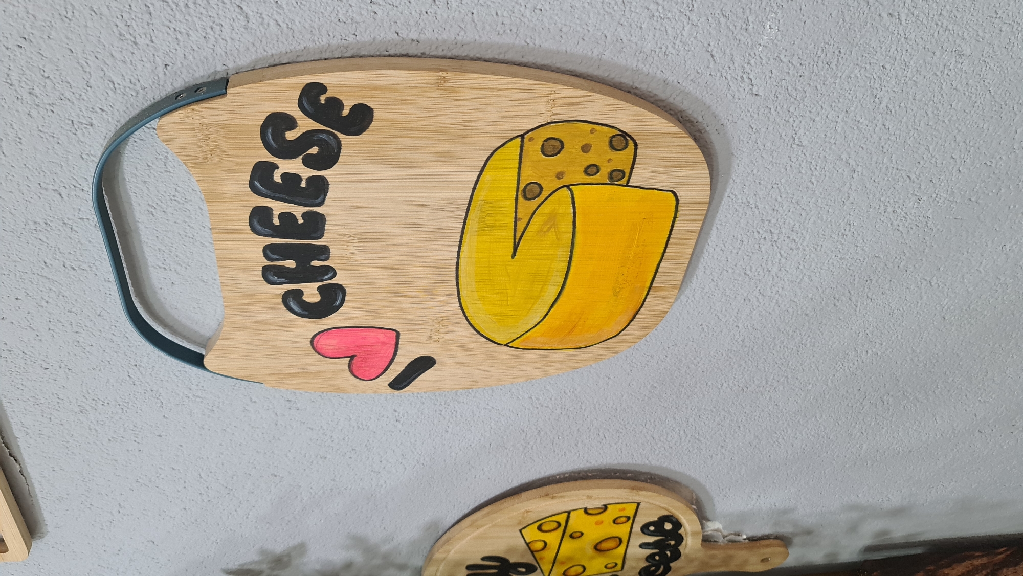A close-up shot shows two cheese-themed wooden signs hanging on a white textured wall. The top sign is a light brown, paddle-shaped cutting board with a turquoise handle. Painted on the board is the word "CHEESE" in thick black letters, followed by a pink heart and a short black line representing the word "I". To the right is a drawing of yellow cheese. A similar, smaller sign is partially visible below, also featuring cheese-related art. The scene does not reveal any location details or contain people.
