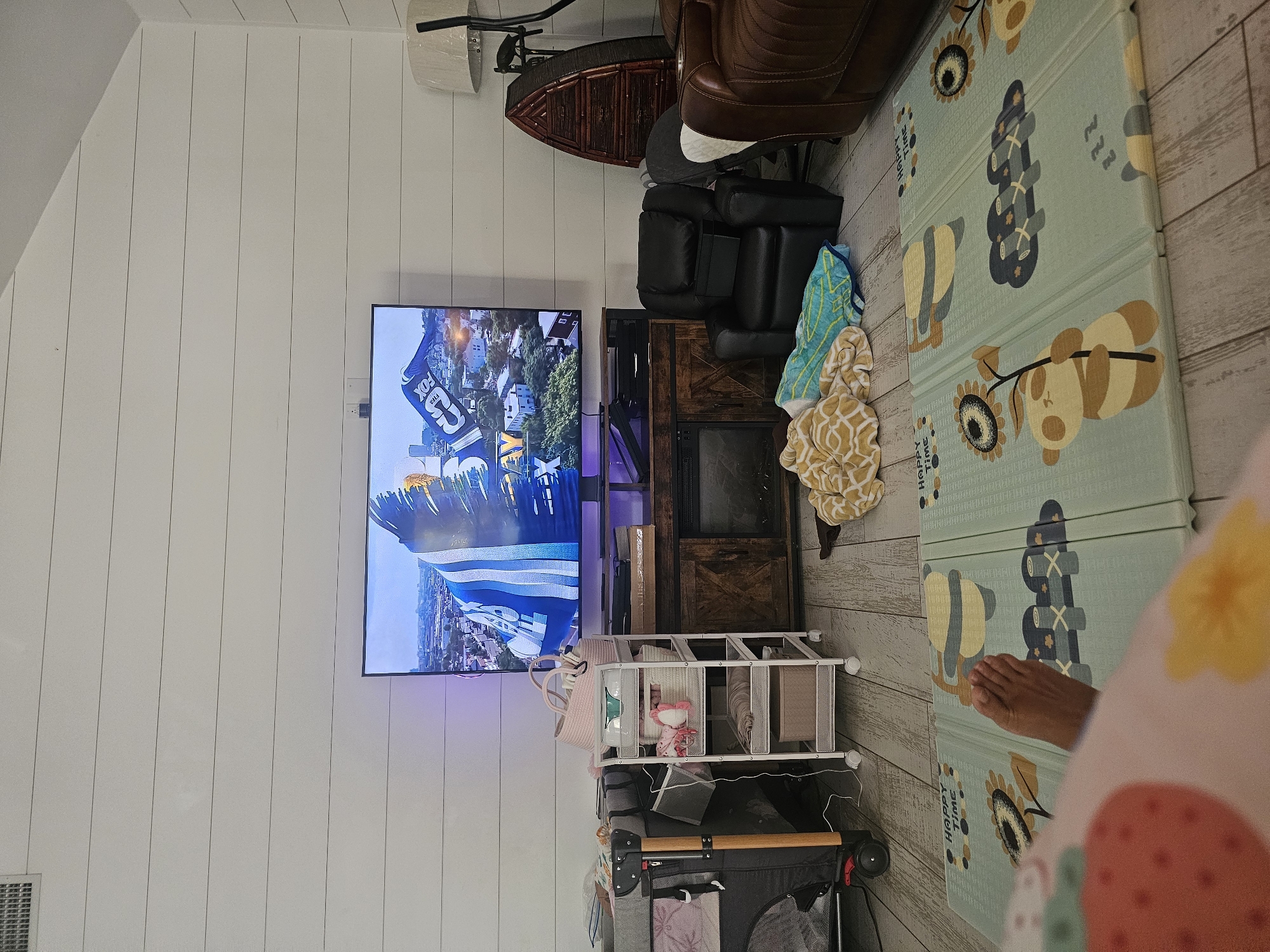 A foot is visible in the foreground of a living room, resting on a light blue playmat featuring cartoon pandas and sunflowers. The playmat is situated on a light wood floor. In the midground, a black recliner chair is positioned next to a brown leather armchair. Behind the recliner, a large flat-screen television displays a sports broadcast, with a man in a blue and white uniform clearly visible. A dark wood media console sits beneath the television. To the left of the television, a white rolling cart is filled with baby items, including pink baskets and blankets, and next to it is a grey and black baby playpen. A tall floor lamp with a white shade stands to the right of the armchair, next to a decorative wooden object resembling a boat. The walls are paneled with horizontal white planks. The scene depicts a home interior, likely in Islamorada, United States of America, set up for relaxation and childcare.