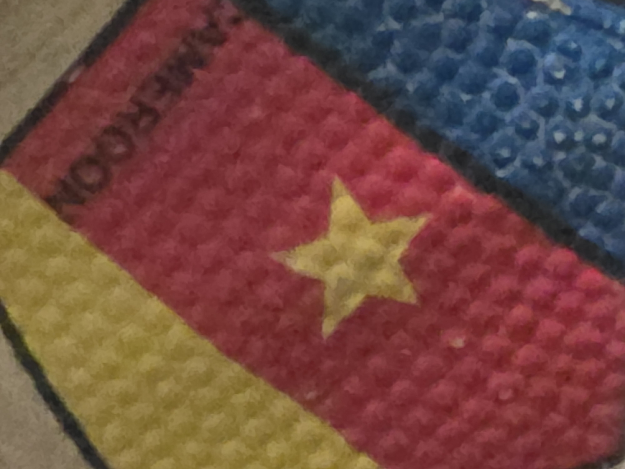 The image shows a close-up of a fabric item with diagonal stripes of yellow, red, and blue. A yellow five-pointed star is centered on the red stripe. The word "AMEROON" is written in black text diagonally across the red stripe. The fabric appears to be textured, possibly knitted or embroidered. The overall color palette is vibrant, suggesting it might be part of a flag, banner, or clothing item. The background is out of focus and appears to be a neutral color. No specific location in a city can be determined from the image.