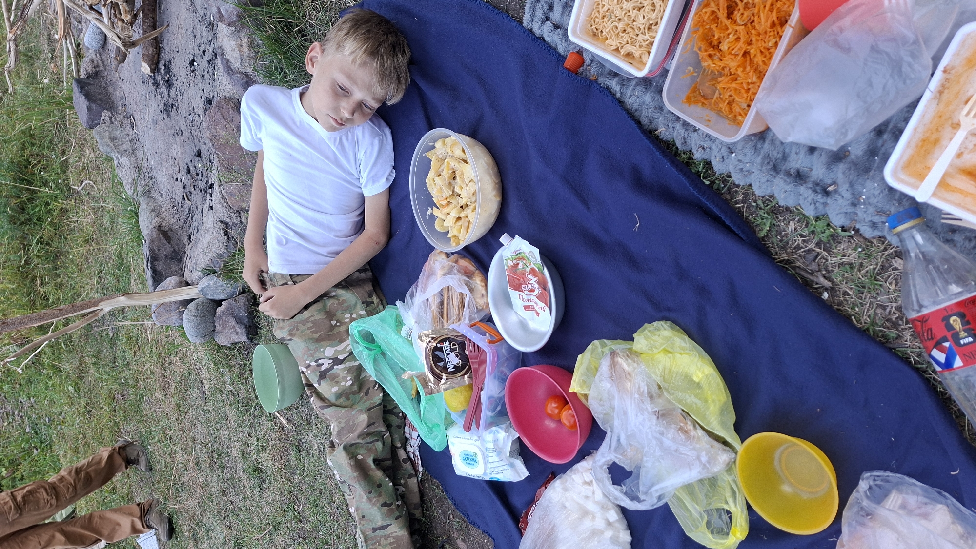 A young boy with blonde hair and blue eyes lies on his back on a blue blanket spread on the ground. He is wearing a white t-shirt and camouflage pants. His head is tilted back and his eyes are open, looking slightly upwards and to the right. Around him are various food items and containers, suggesting a picnic or camping meal. There are plastic bags filled with food, bowls containing what appears to be pasta salad, tomatoes, and shredded carrots, as well as a bottle of soda. In the background, a person's legs, wearing brown pants and hiking boots, are visible, as is a campfire pit made of stones. The ground is covered with grass and dirt. The scene appears to be outdoors, possibly in a natural setting near a campsite. There is no specific indication that the image was taken in the city of Batan, Kazakhstan.