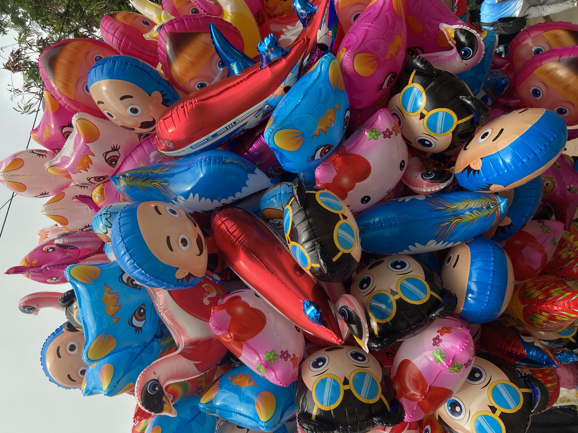 A dense cluster of inflated foil balloons fills the frame, their reflective surfaces catching the light. The balloons depict various cartoon characters and designs, including stylized human faces with blue hair and mustaches, aviator-style faces in black with blue-tinted goggles, pink cat-like faces with flowers, and blue bird-like faces. Several long, red, blimp-shaped balloons are interwoven amongst the character balloons. The balloons are likely being held or sold, as there is an indistinct, bright sky and blurred green foliage visible in the top portion of the image, implying an outdoor setting. The scene could be taking place in Dhaka, Bangladesh, but there is no visual confirmation within the image to confirm that location.