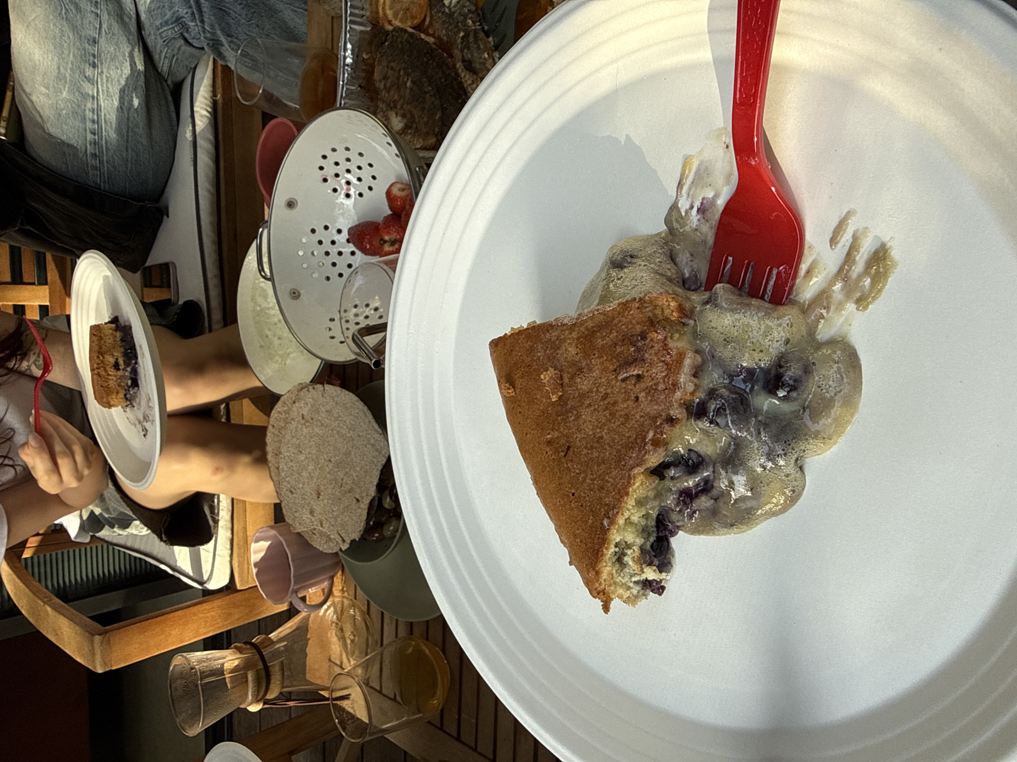A bird's-eye view captures a casual outdoor dining scene, likely on a patio or balcony, with a group of people enjoying a meal.
The main focal point is a white plate displaying a slice of cake covered with a glaze of blueberries and sauce. A red plastic fork is inserted into the cake, suggesting someone is in the process of eating.
To the left of the plate, a person is holding a smaller white plate with another piece of similar cake. Various items are scattered around the table, including a colander with strawberries, a coffee pour-over setup, mugs, and food in containers.
The table appears to be made of wooden slats, typical of outdoor furniture. A person wearing denim jeans is seated next to the table. The setting suggests a relaxed atmosphere, perhaps a brunch or afternoon gathering. There is no verifiable information to ascertain the location.