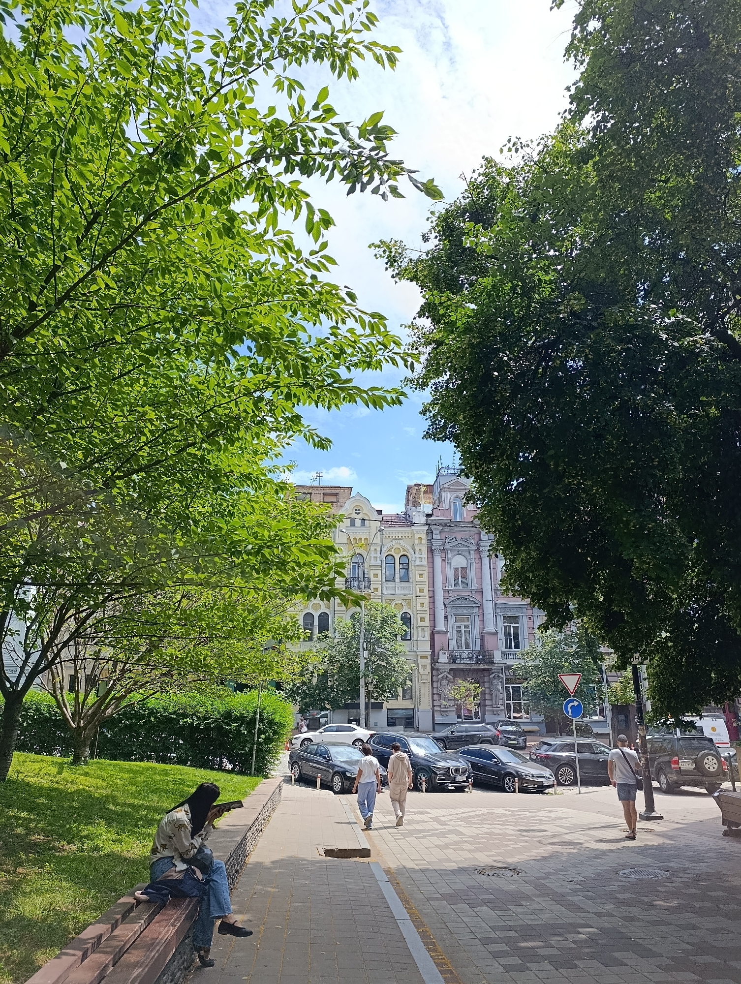 In Kyiv, Ukraine, on a sunny day, the scene unfolds on a tree-lined street. In the foreground, a person sits on a wooden bench with their back to the viewer, engrossed in a book or phone, with a backpack beside them. Green grass and a hedge form the backdrop to the bench. A paved walkway runs alongside the street, where several cars are parked. Behind the parked cars, ornate, multi-story buildings with distinct architectural details and balconies line the street. Two people walk on the walkway, one in a light-colored outfit and the other in a white top and blue pants. Lush green trees with dense foliage frame the scene, with branches reaching towards a partly cloudy blue sky. A street sign with a triangular warning and a circular blue sign indicating a direction are visible.