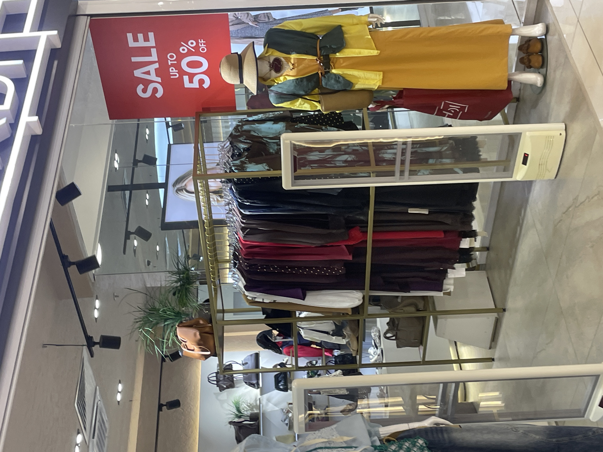 An indoor retail store display shows a mannequin dressed in a yellow long-sleeved shirt, a dark green vest, and a long yellow skirt. The mannequin wears a wide-brimmed hat, a necklace, and a brown belt. A brown purse is slung over its shoulder. The mannequin stands on a circular platform with two brown shoes. To the left of the mannequin is a rack of clothing on hangers, featuring various colors including purple, burgundy, and red. A red shopping bag with white lettering is behind the mannequin. A security gate with a red light is positioned in front of the clothing rack. In the background, shelves are stocked with handbags and decorative plants. A large red sign with white text reading "SALE UP TO 50% OFF" is visible. The scene suggests a commercial setting, likely a clothing boutique in Tashkent, Uzbekistan.